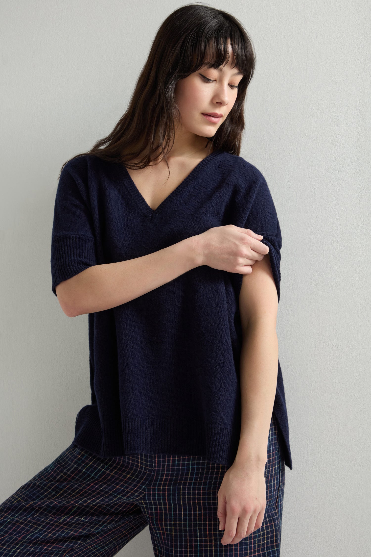 A woman with long hair wears the Merino V Neck Knot Stitch Jumper in navy and plaid pants as she looks down adjusting her sleeve, showcasing the beautifully draped merino wool fabric with subtle knot stitch detailing.
