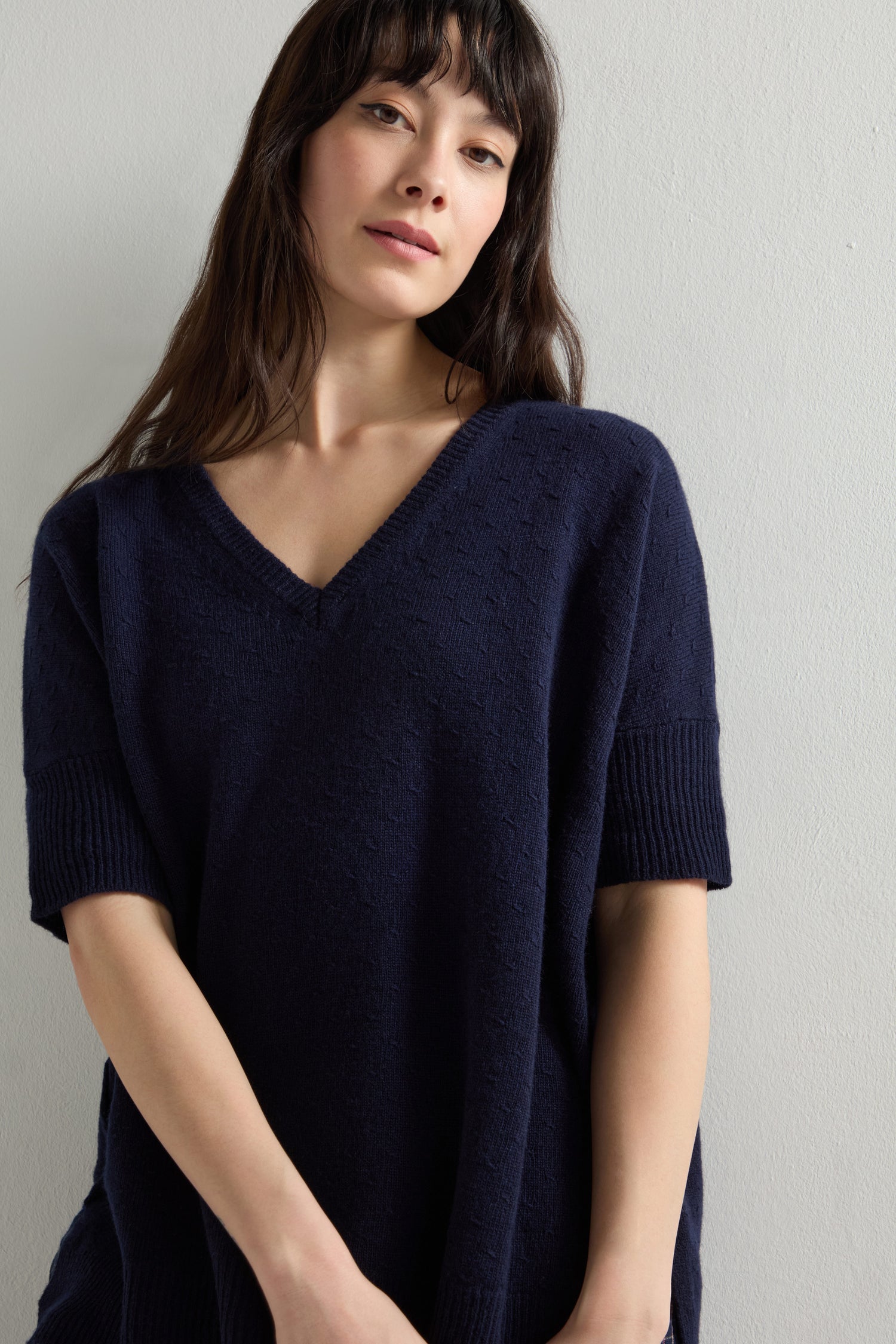 A person with long dark hair stands against a white wall, wearing a Merino V Neck Knot Stitch Jumper in navy, crafted from soft Merino wool.