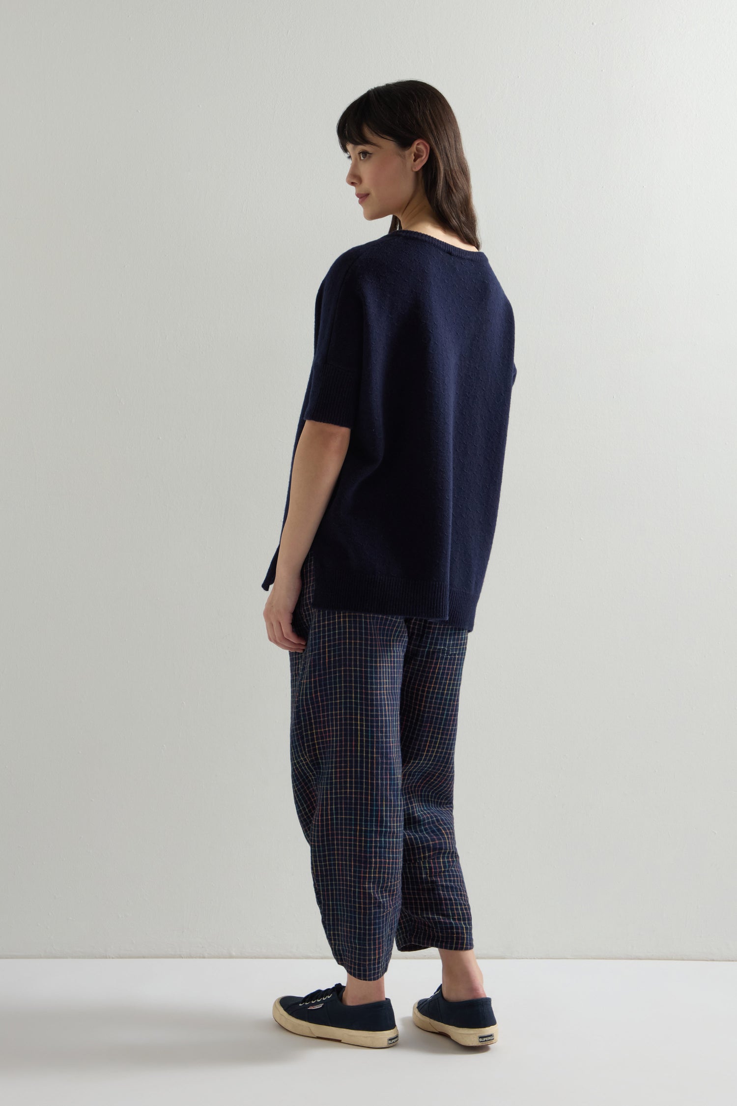 A person standing sideways in a Merino V Neck Knot Stitch Jumper in dark blue, paired with patterned pants and dark sneakers, against a plain white background.