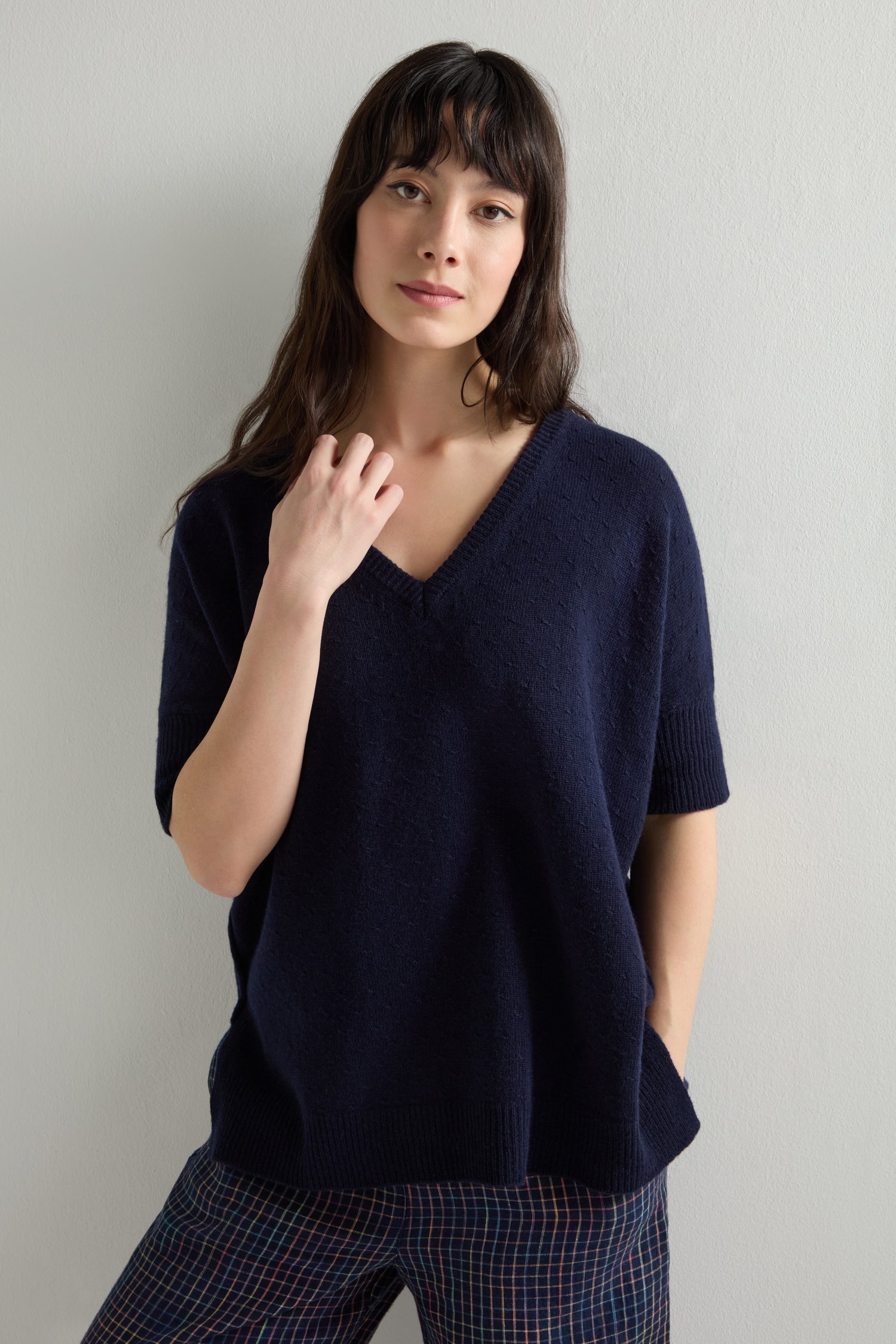 A person with long hair stands against a plain backdrop, wearing a Merino V Neck Knot Stitch Jumper in dark blue and plaid pants.