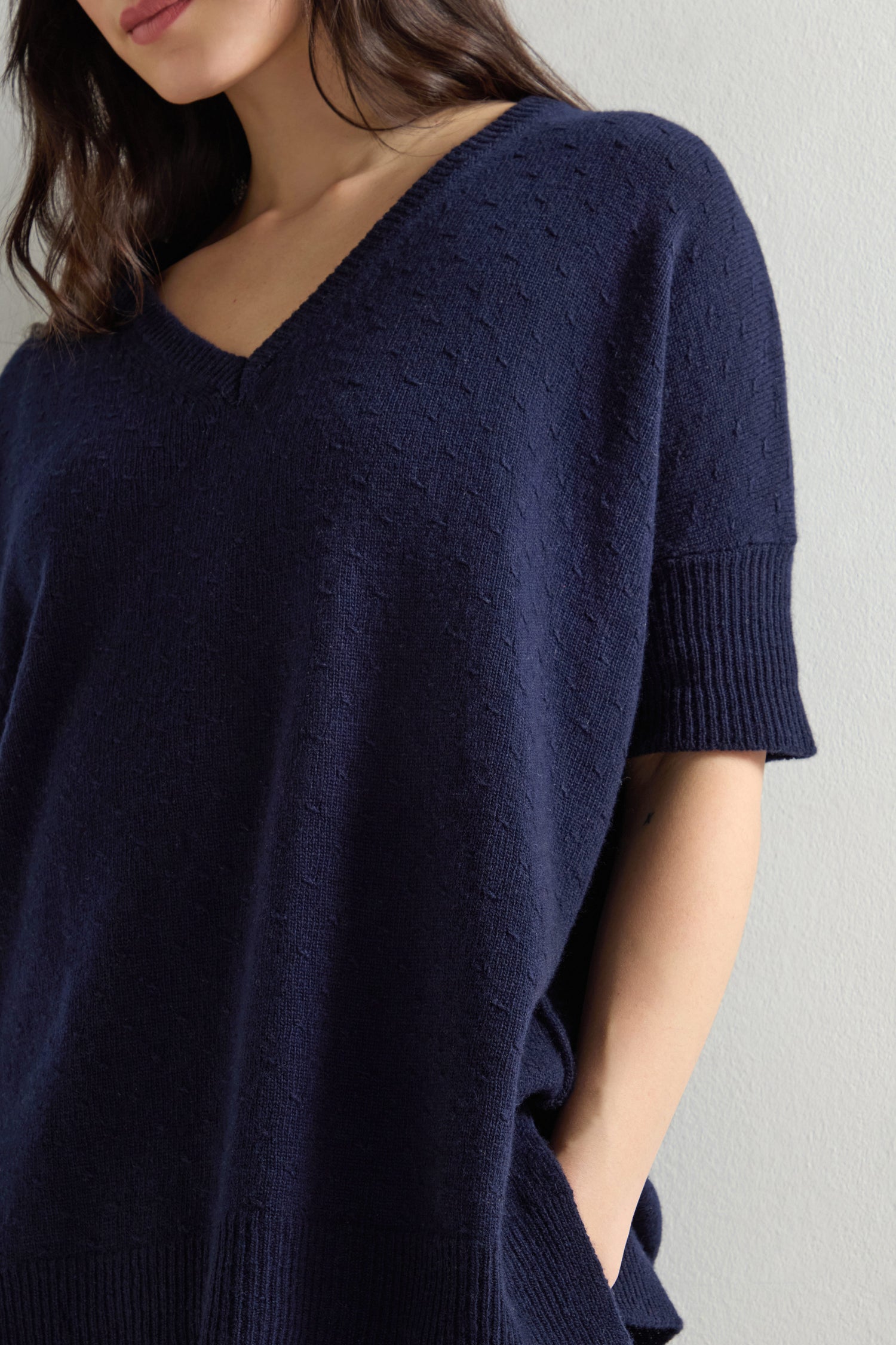 A person stands against a neutral background, exuding elegance and understated charm in a Merino V Neck Knot Stitch Jumper in dark blue.