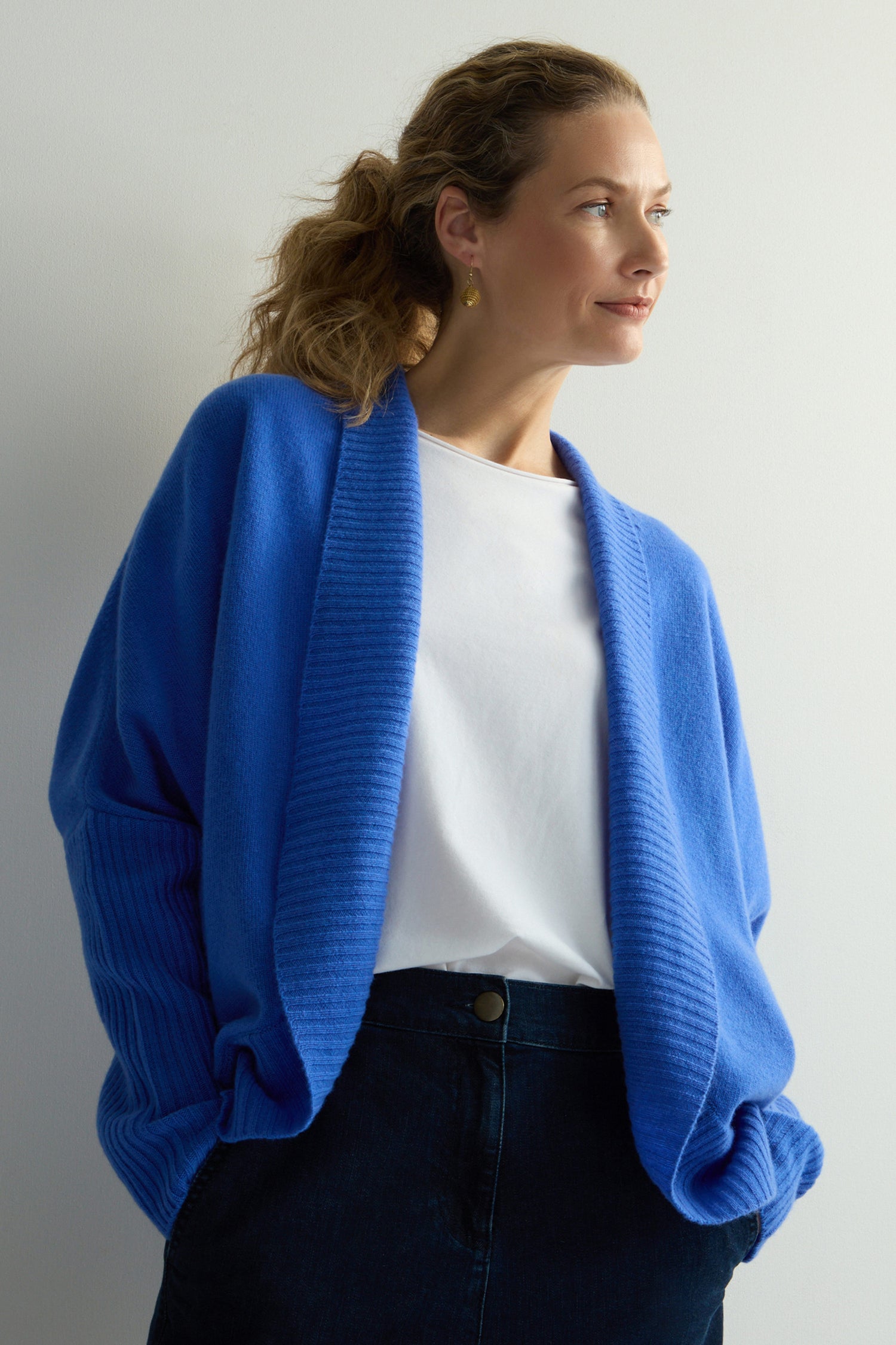 The person wears a calming blue Merino Ribbed Cuff Shawl Cardigan, elegantly paired with a white shirt and dark pants, looking to the side.