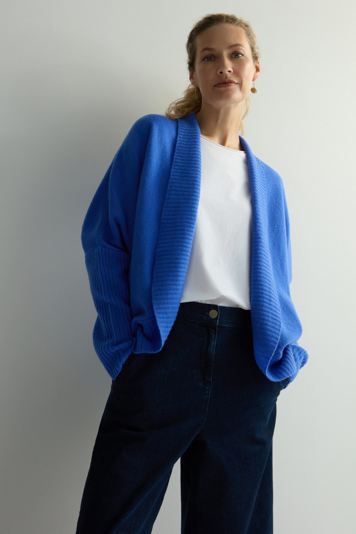 A person stands against a plain background, hands in pockets, wearing a Merino Ribbed Cuff Shawl Cardigan from the knitwear collection, along with a white shirt and dark pants.
