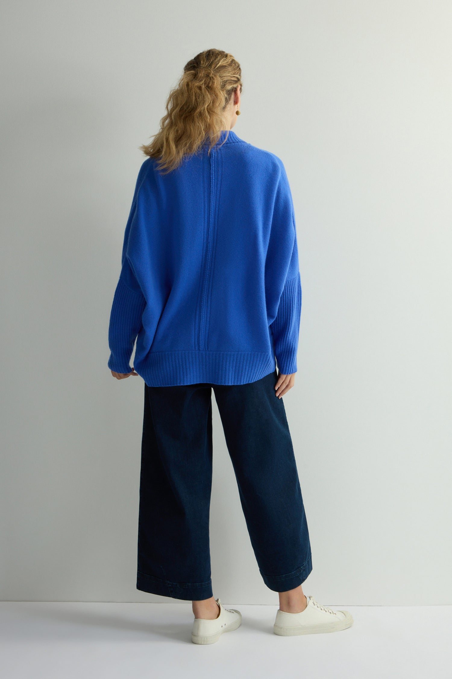 A woman with wavy hair, facing away, models a bright blue Merino Ribbed Cuff Shawl Cardigan with dark wide-leg pants and white sneakers against a plain white background.