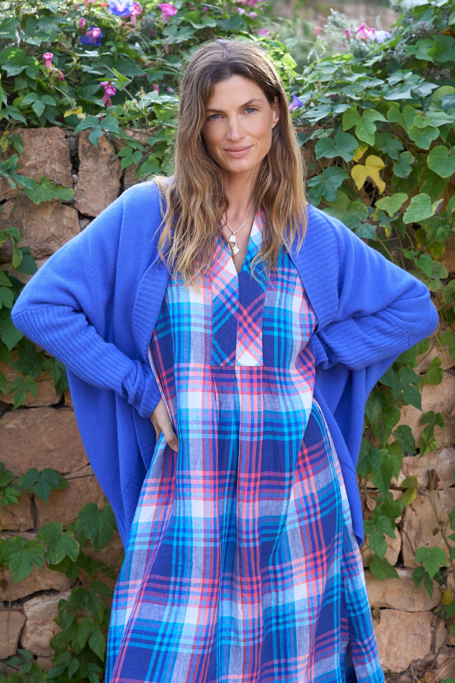 A woman with long hair wears a Merino Ribbed Cuff Shawl Cardigan over a plaid dress, standing before a stone wall decorated with green foliage and flowers.