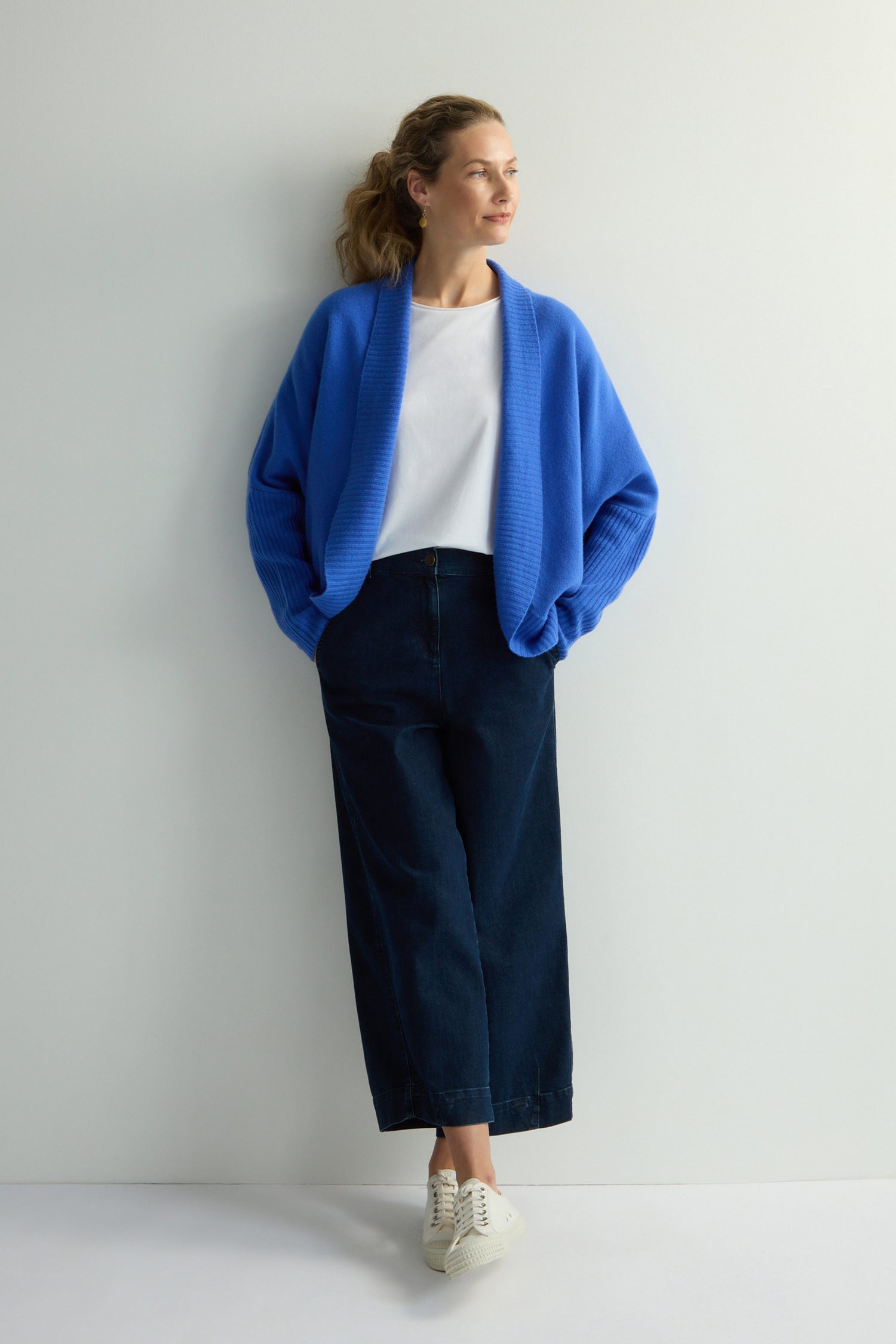A person in a Merino Ribbed Cuff Shawl Cardigan from the knitwear collection, paired with a white shirt and dark wide-leg pants, leans against a white wall with hands in pockets, wearing white sneakers.