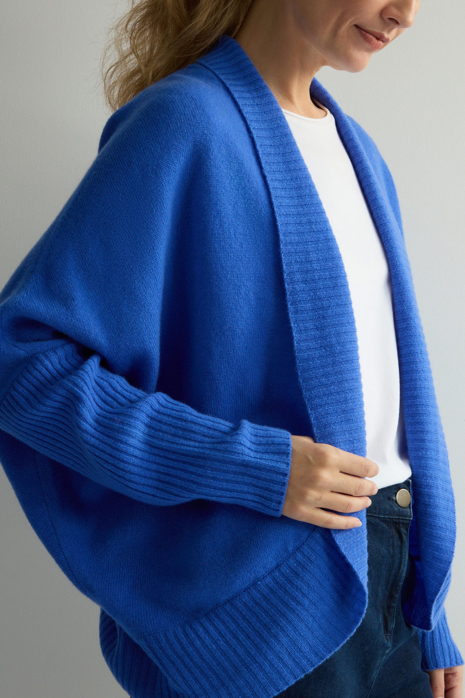 A person in a blue Merino Ribbed Cuff Shawl Cardigan from the Knitwear Collection, layered over a white shirt and jeans, stands sideways.