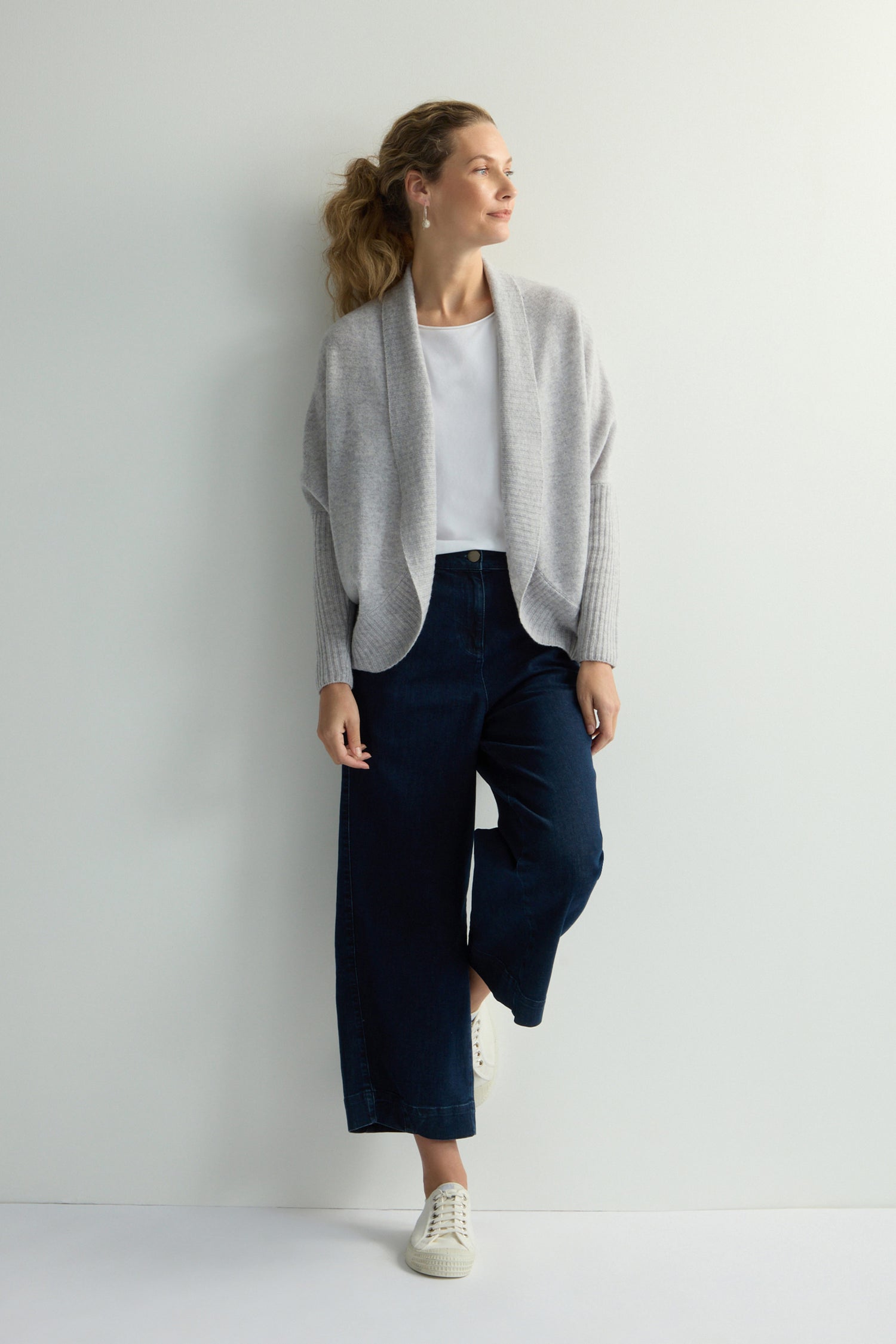 A person wearing the Merino Ribbed Cuff Shawl Cardigan in gray, paired with a white shirt, blue wide-leg jeans, and white sneakers from the knitwear collection, stands against a plain white wall.