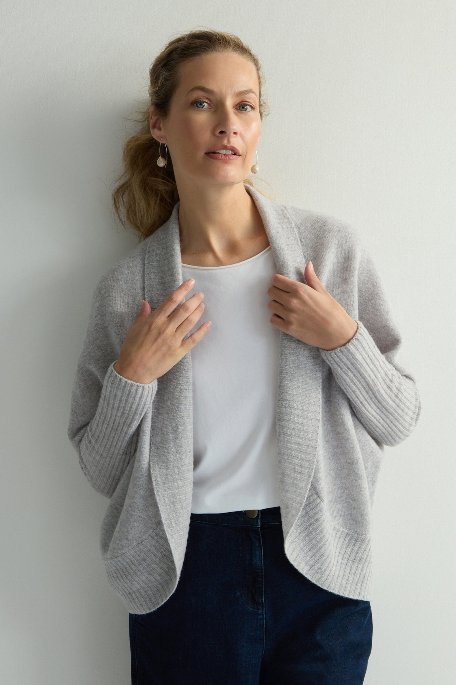 A woman in a light gray Merino Ribbed Cuff Shawl Cardigan and white shirt stands against a plain background, her hands gracefully near the cardigan's collar.