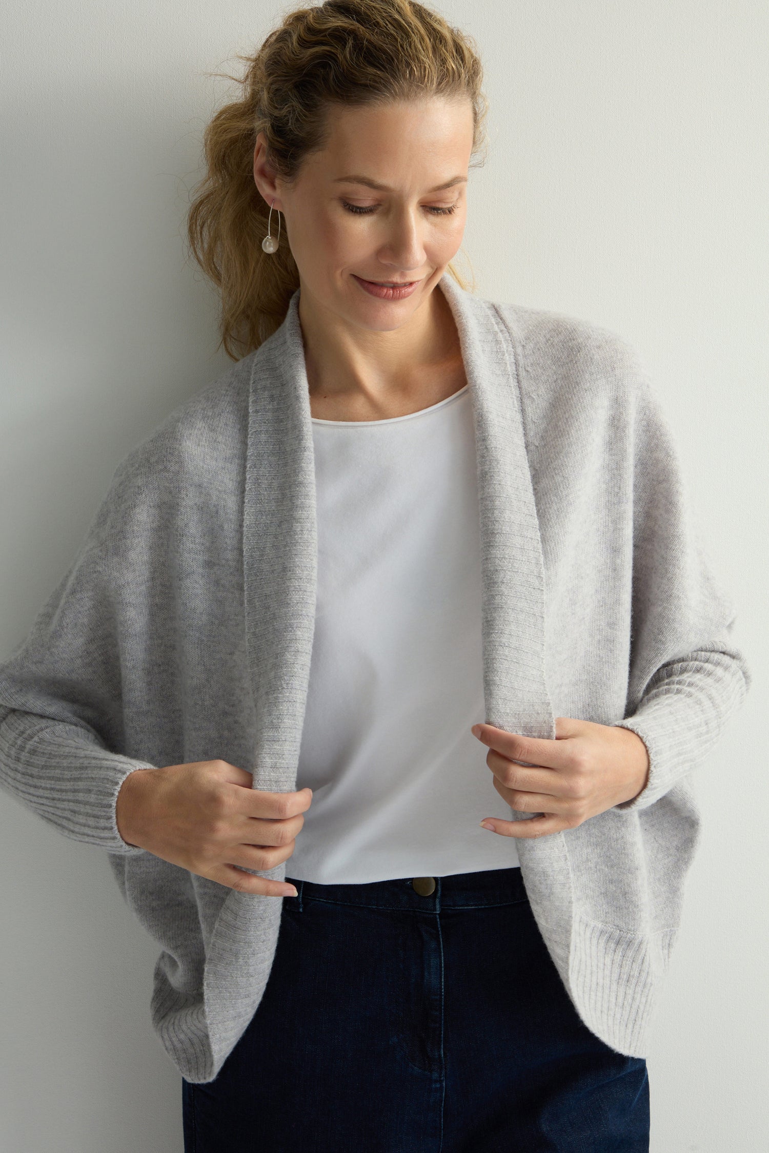 A person adjusts a gray Merino Ribbed Cuff Shawl Cardigan over a white shirt, standing against a plain background, looking down, showcasing the new Knitwear Collection.
