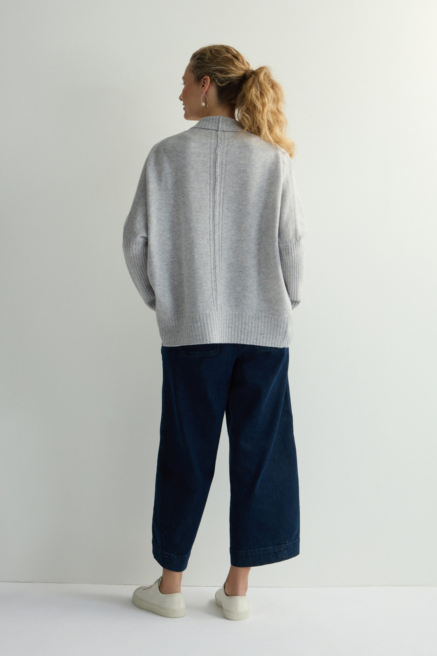 A person with light-colored hair, seen from behind against a plain background, is wearing a gray Merino Ribbed Cuff Shawl Cardigan, dark wide-leg pants, and white sneakers.