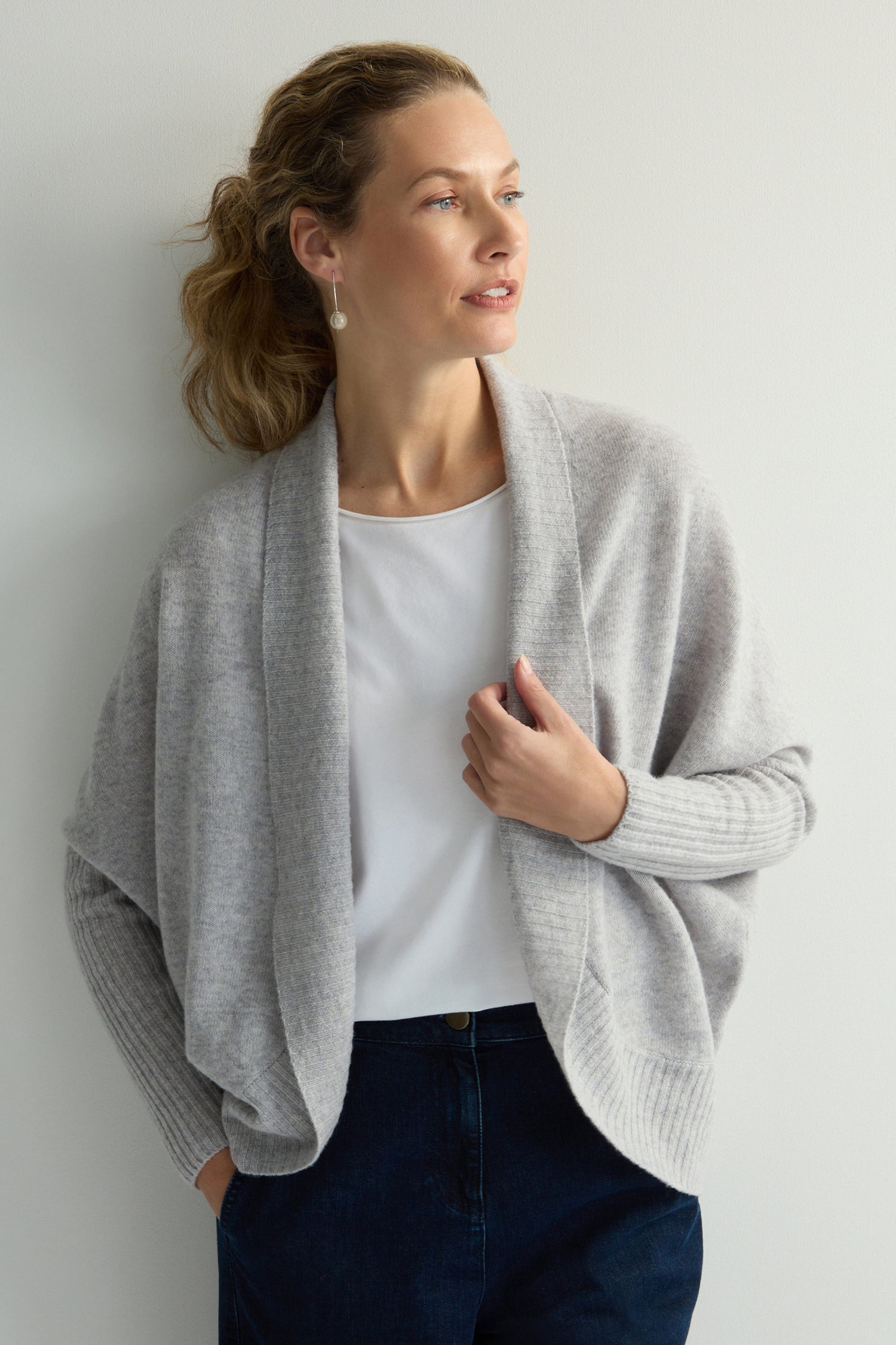 A woman in a gray Merino Ribbed Cuff Shawl Cardigan and white shirt stands against a light background, looking to the side with one hand on her knitwear.