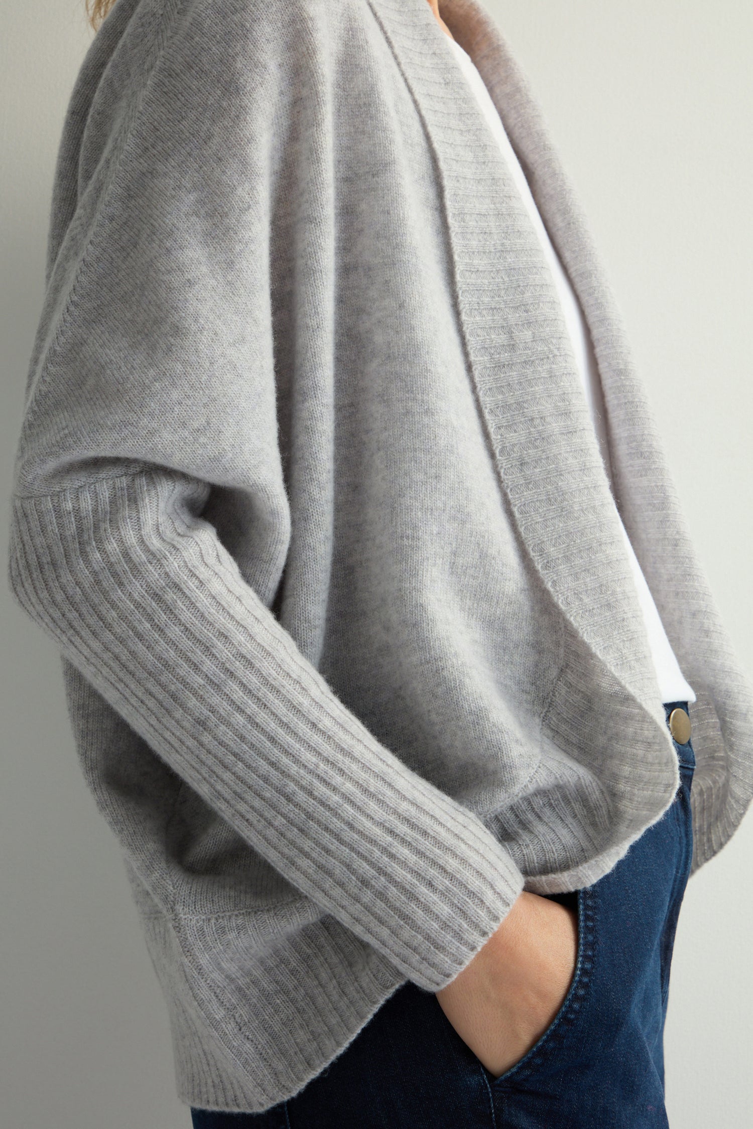 A person wears a Merino Ribbed Cuff Shawl Cardigan over a white shirt and blue jeans, with hands tucked comfortably in their pockets.