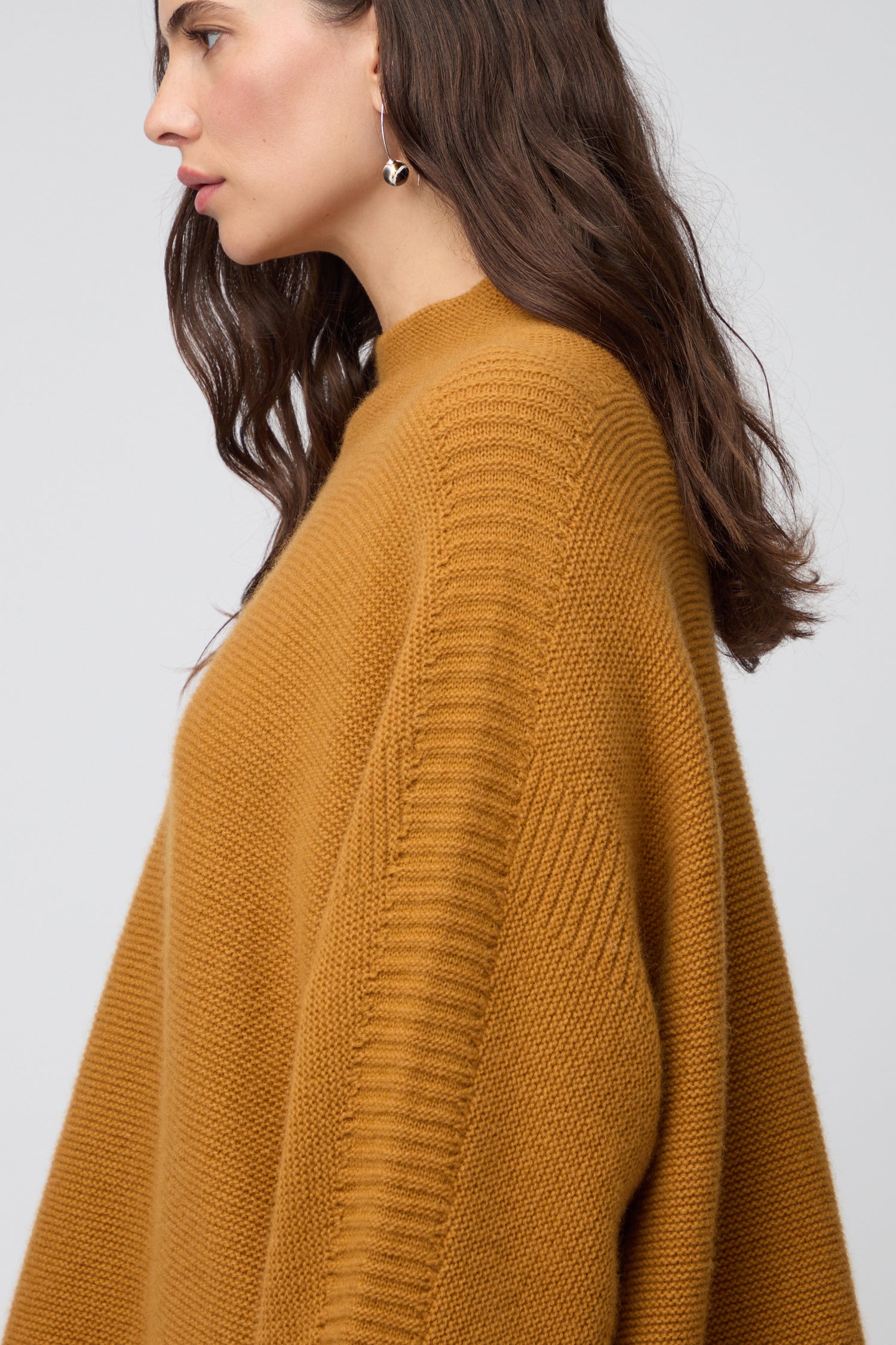Person wearing the Merino Wool Stitch Detail Jumper in textured mustard yellow, featuring a relaxed boxy silhouette and side slit, paired with dark pants; photo highlights the lower half of this knitwear staple and part of their arm.