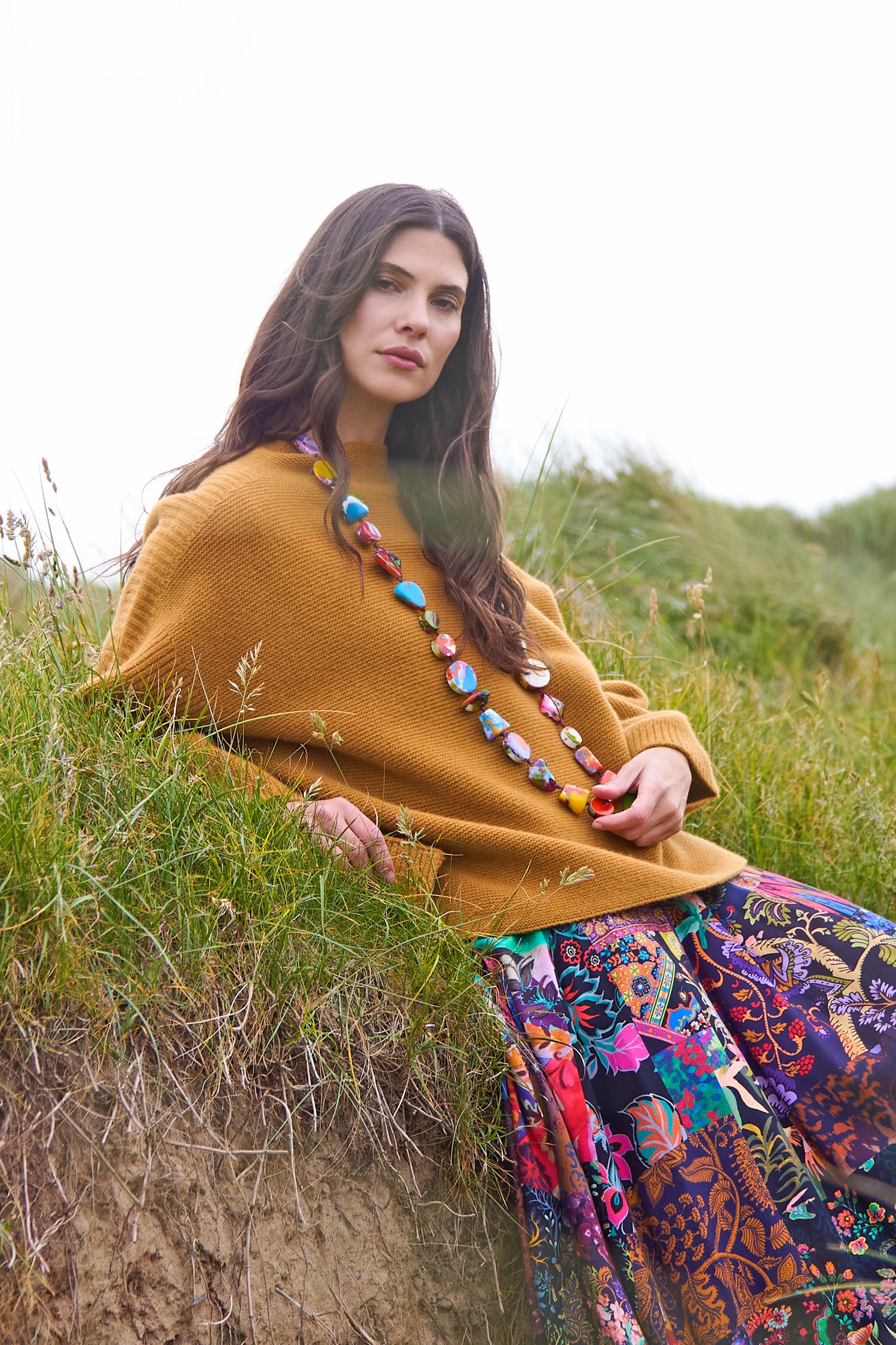 A woman with long dark hair sits on a grassy hillside, wearing the Merino Wool Stitch Detail Jumper in mustard, a colorful beaded necklace, and a vibrant floral-patterned skirt.