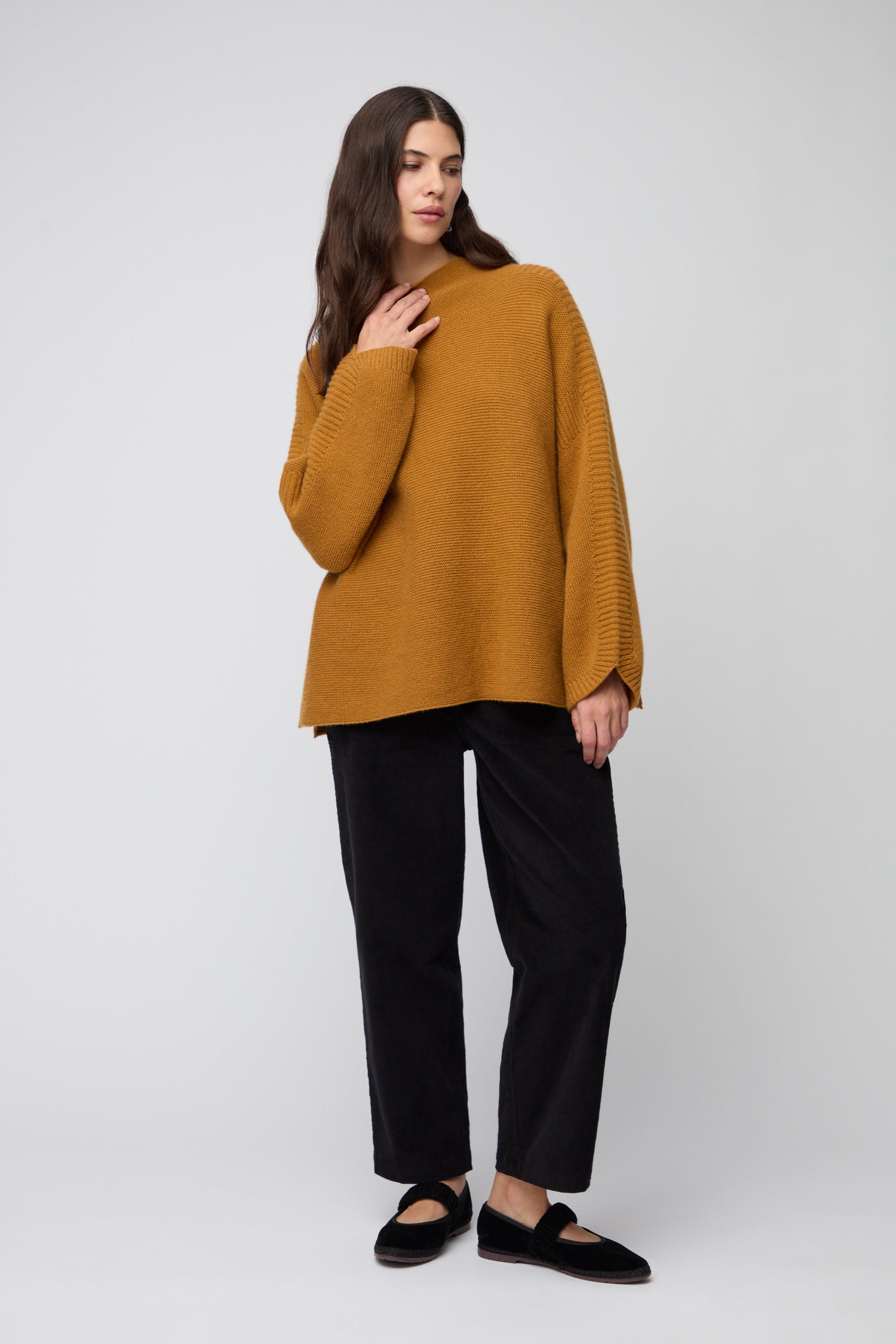 A woman with long dark hair wears the Merino Wool Stitch Detail Jumper in ochre, paired with orange earrings. She looks at the camera, one hand on her collar, highlighting the jumper’s relaxed boxy silhouette against a neutral background.