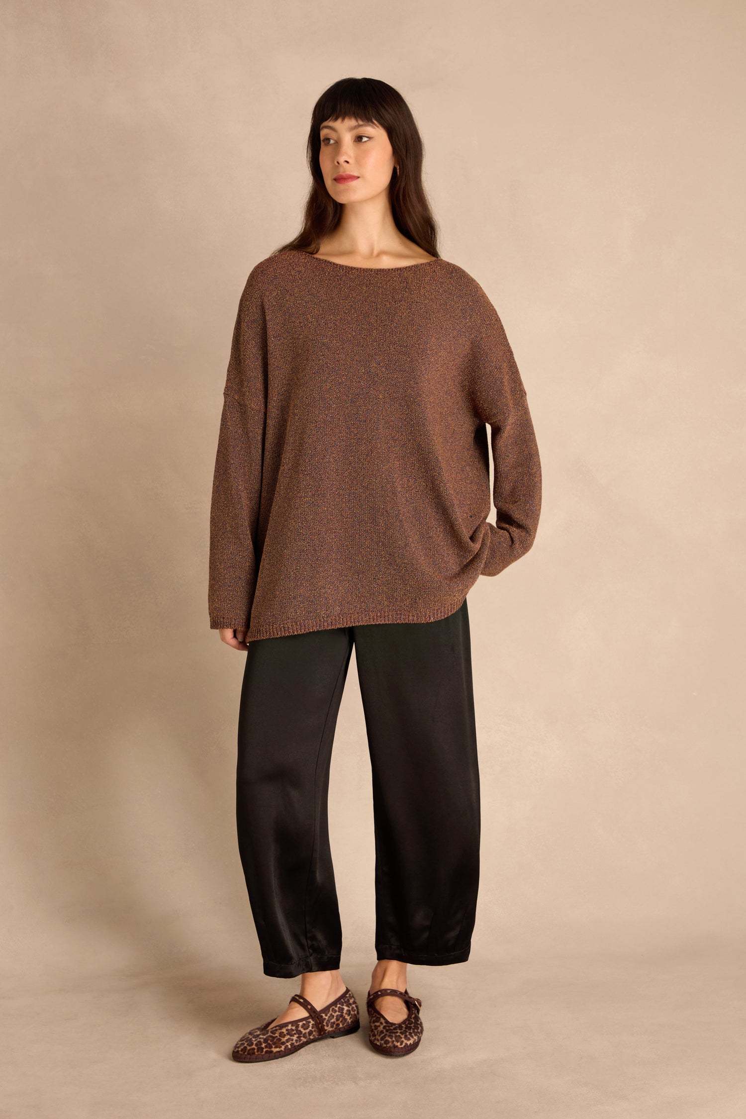 Woman stands against a beige background wearing the Copper Boxy Sparkle Knit Jumper, black wide-leg pants, and leopard print flat shoes.