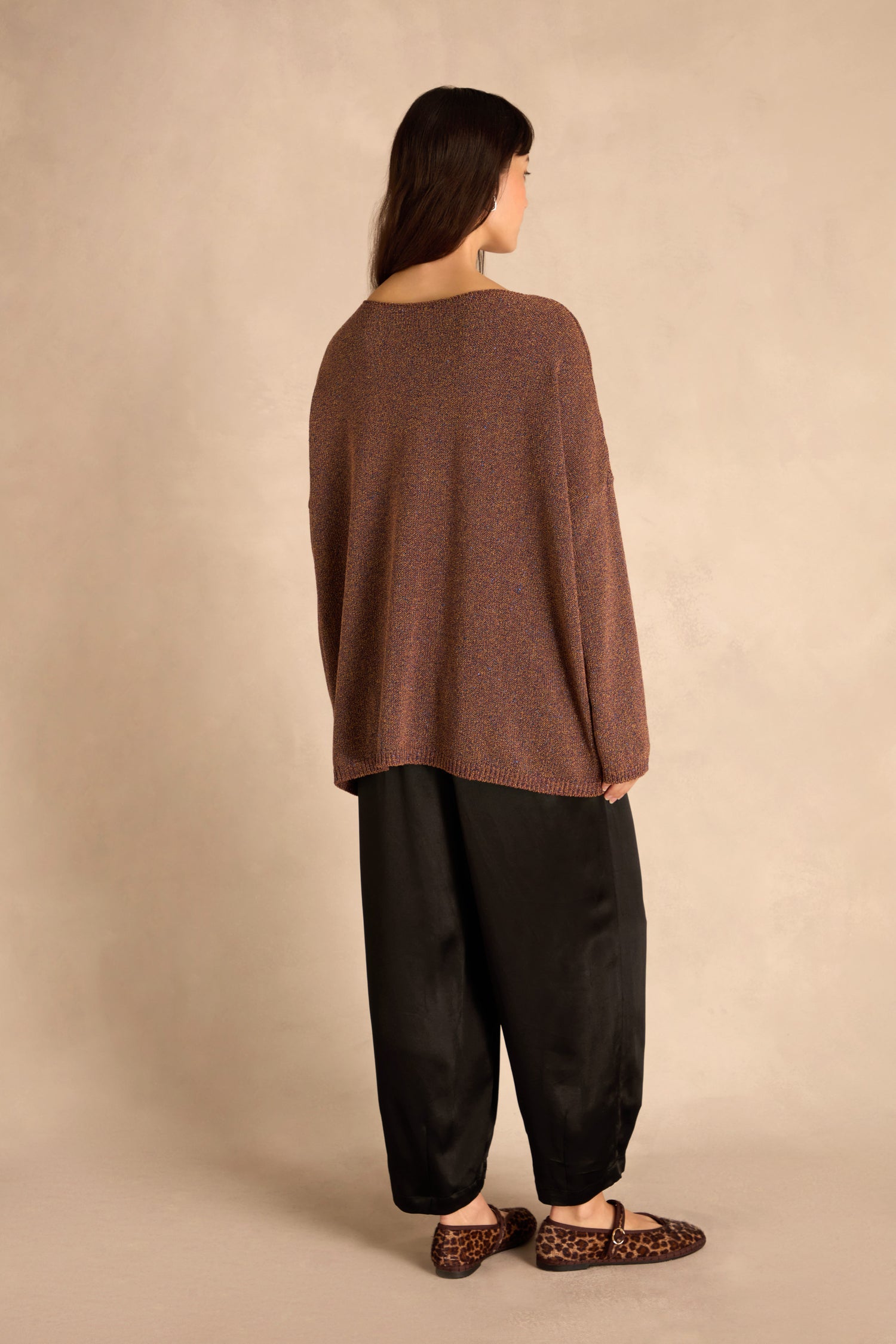 Person stands with back to camera in a boxy silhouette: Copper Boxy Sparkle Knit Jumper, black wide-leg pants, leopard print flats, beige background.