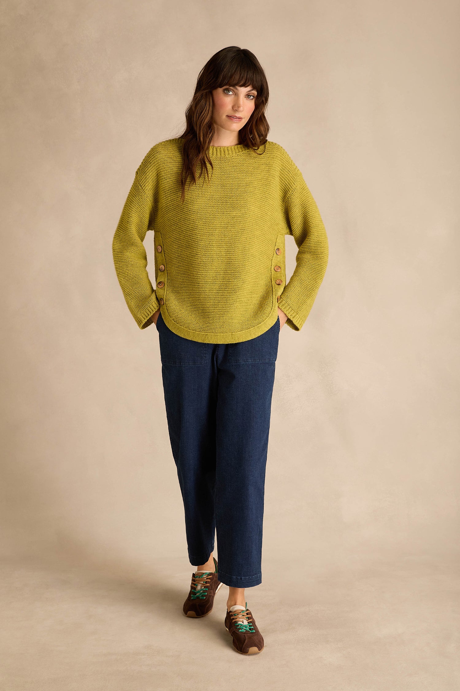 Woman wearing an Avocado Ribbed Wool Blend Button Jumper with blue cropped pants and brown shoes, standing against a beige background.