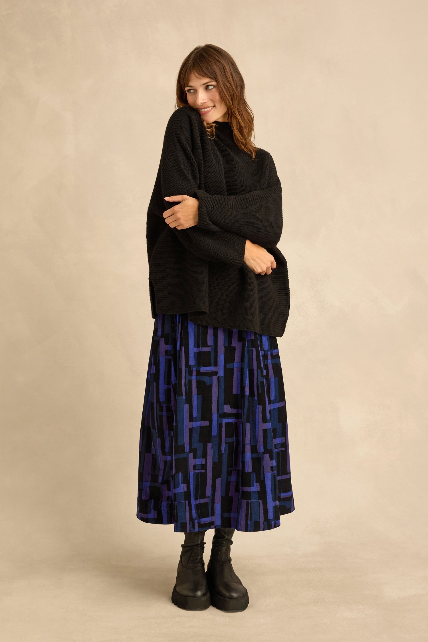 A woman stands against a neutral background, looking to the side and smiling, dressed in a Black Merino Ribbed Turtle Neck Jumper, a blue and black patterned midi skirt, and black boots.