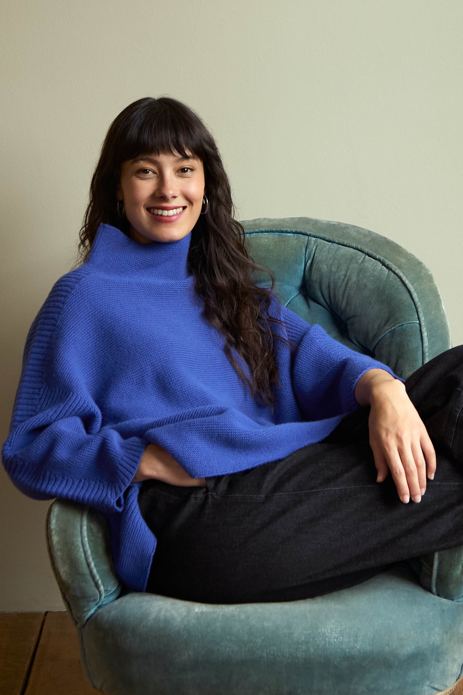 A woman wearing a Royal Blue Merino Ribbed Turtle Neck Jumper sits in a velvet armchair.