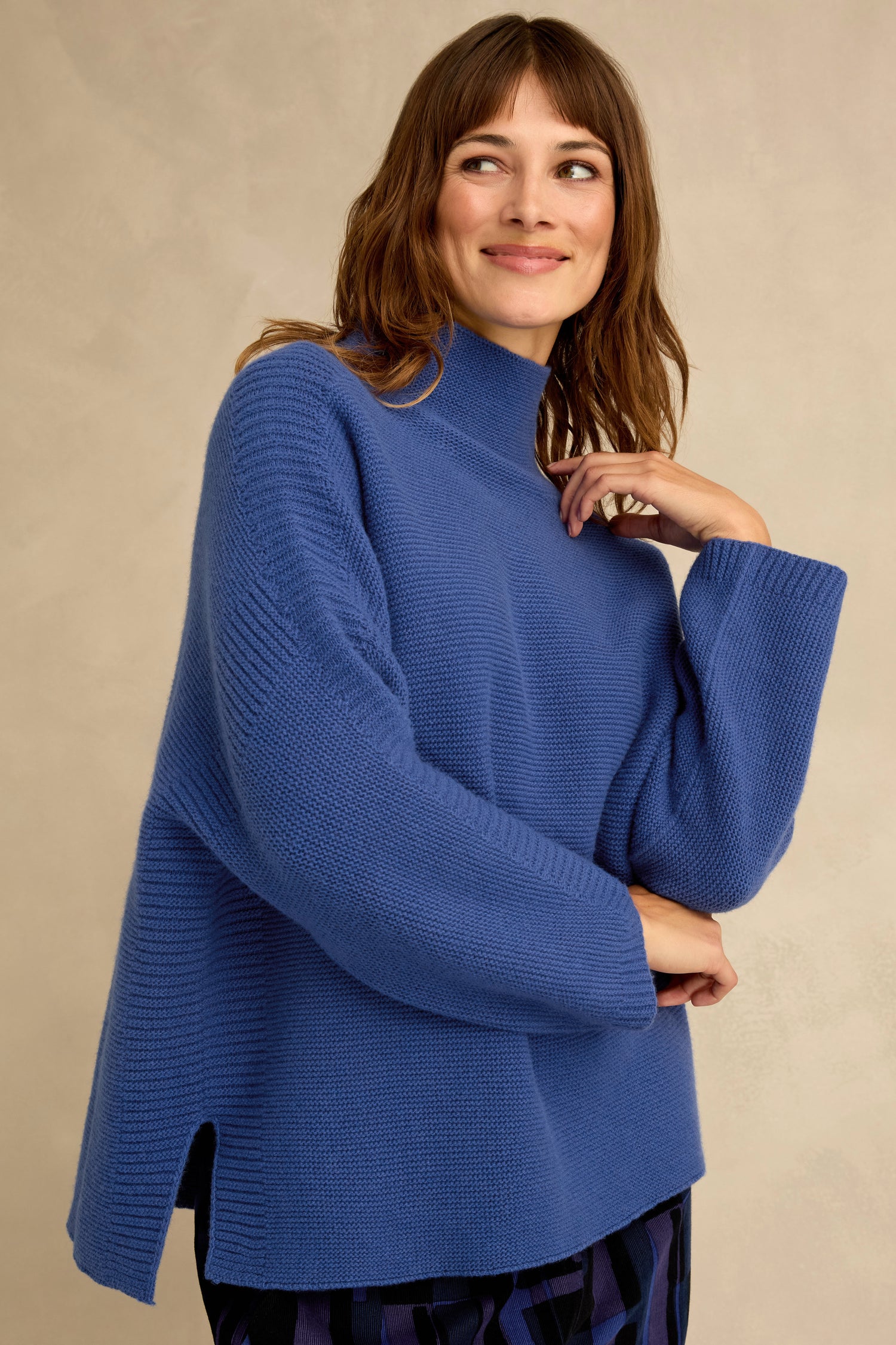 A woman wearing the Royal Blue Merino Ribbed Turtle Neck Jumper, featuring a relaxed fit with side slits and a high neckline, stands against a plain light beige background, smiling and gazing to the side.