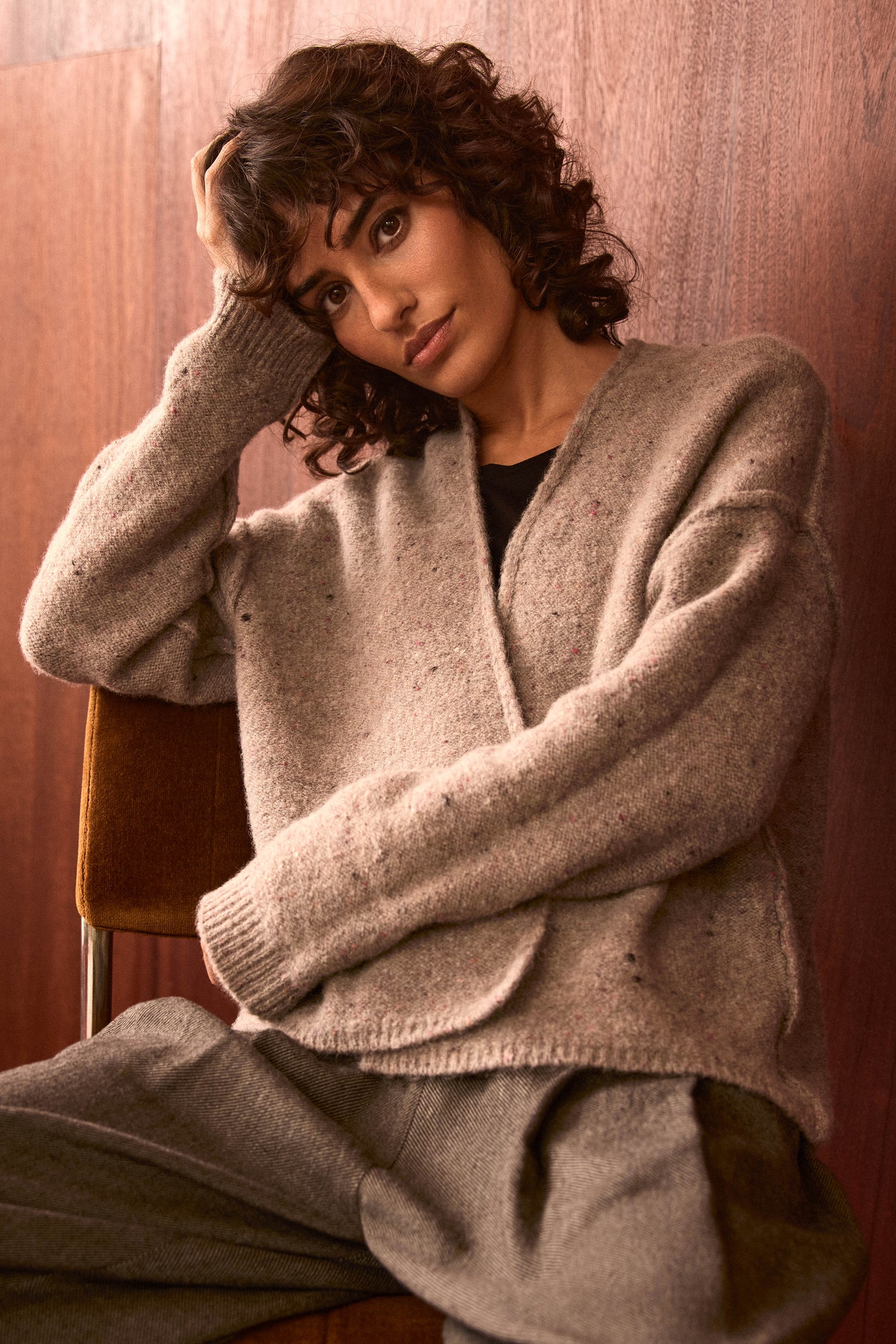A woman with short curly hair sits on a chair, leaning her head on one hand. She wears the Taupe Alpaca Blend Cropped Cardi and gray pants, posed against a wood-paneled wall. This relaxed look blends comfort and effortless style.