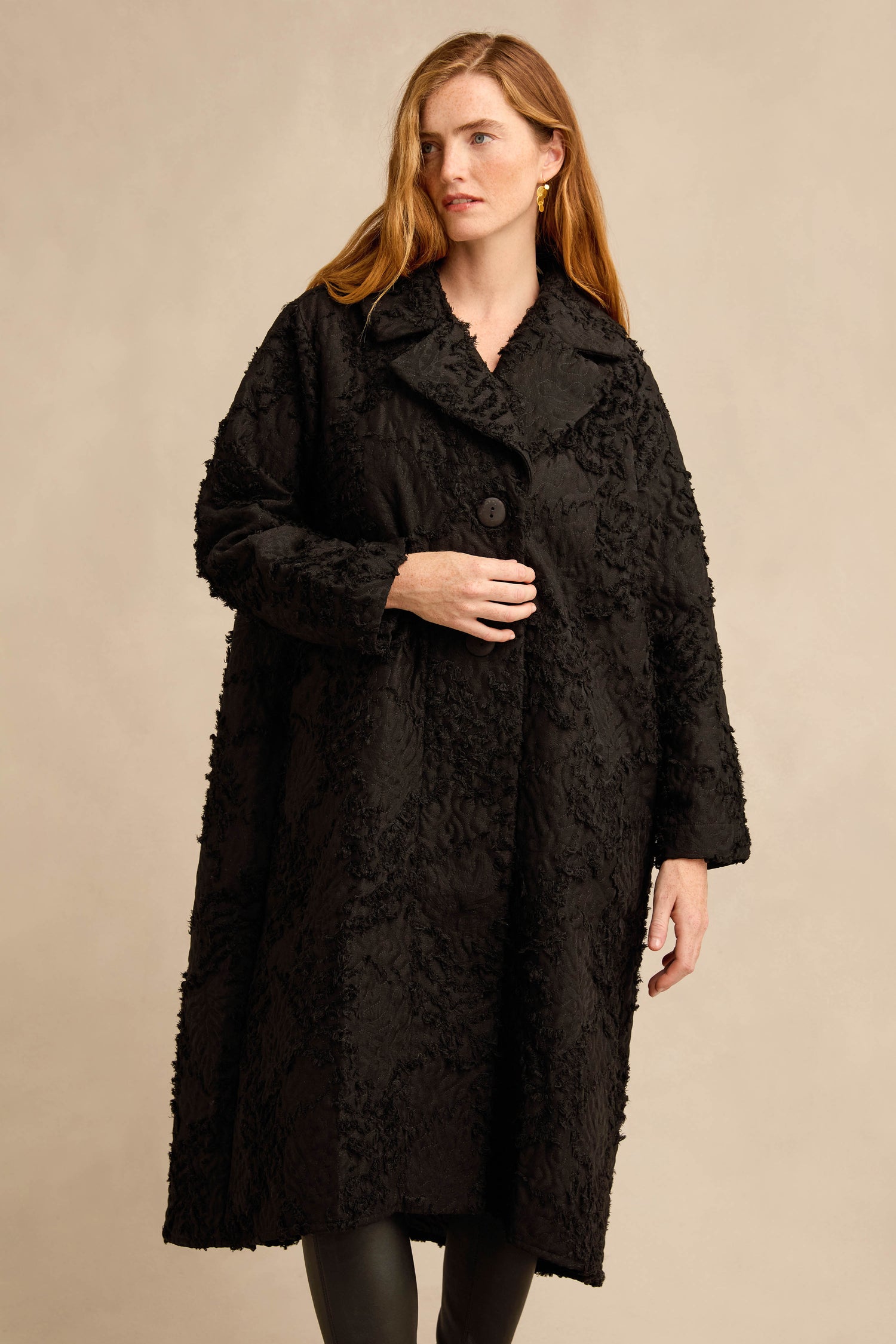A woman wearing the Textural Big Collar Coat—a black, longline piece with long sleeves—standing against a plain beige background.