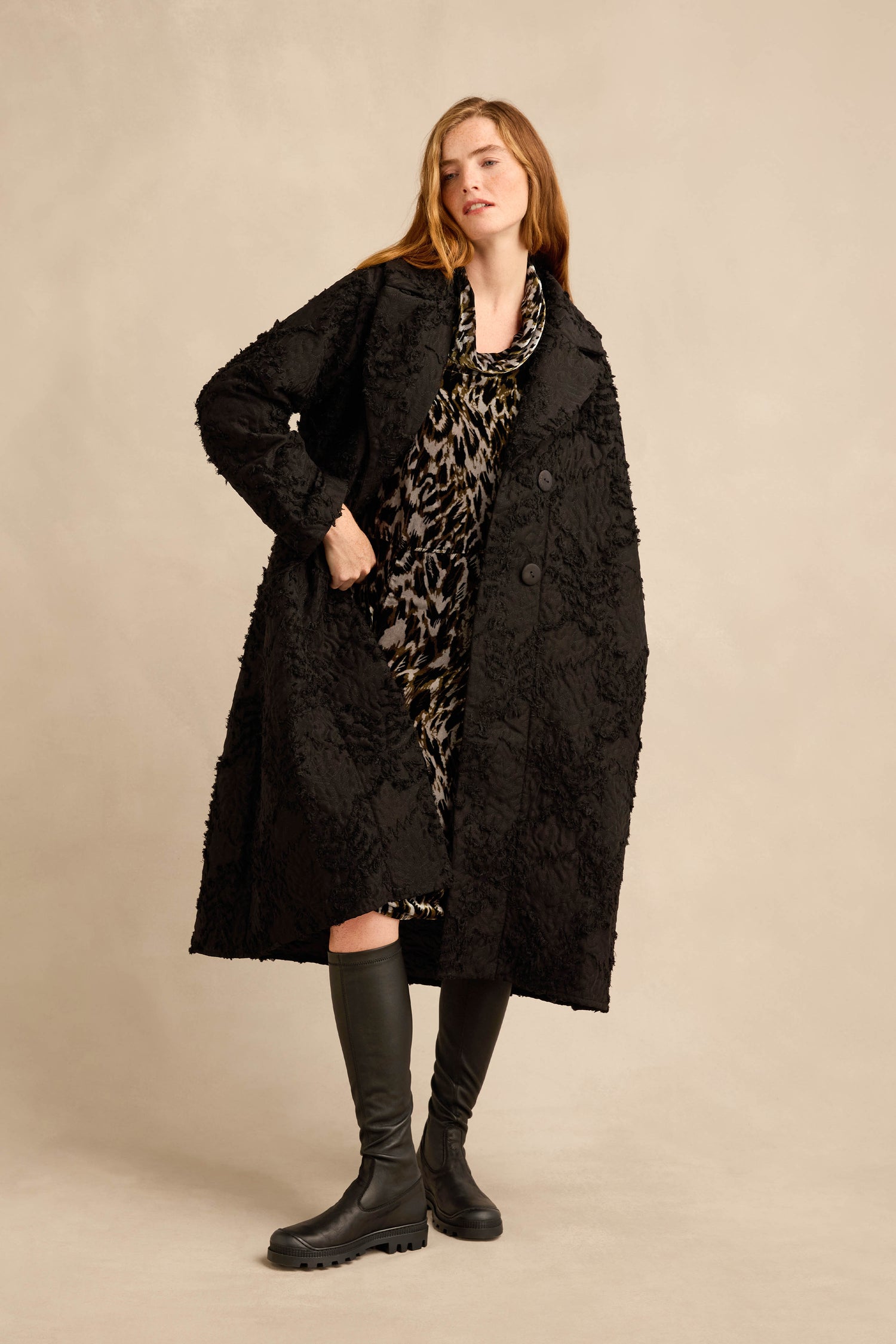 A woman wearing the Textural Big Collar Coat in black over a leopard print dress and black knee-high boots, standing against a beige background.