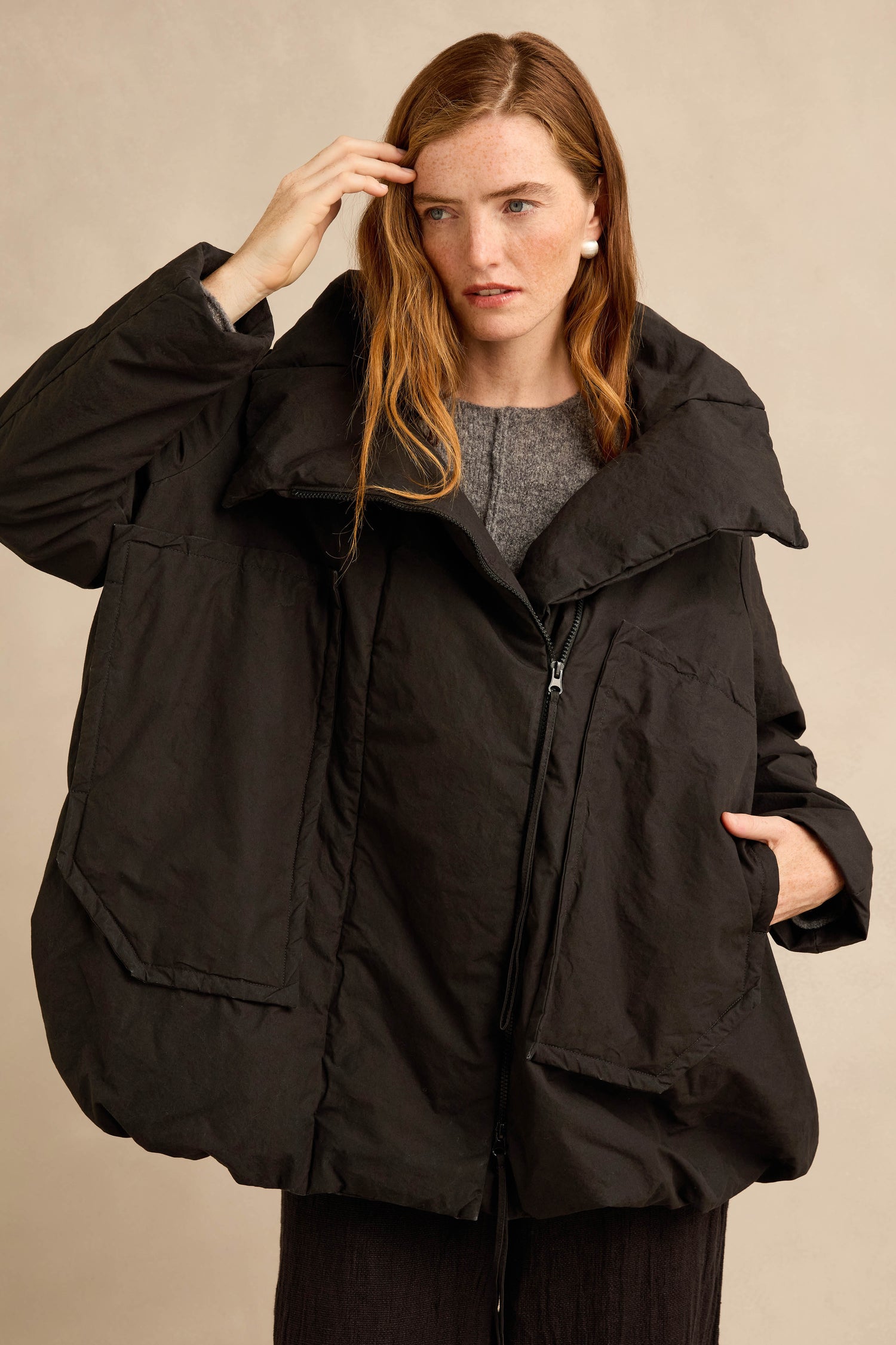 A woman with long red hair wears the Big Pocket Puffer Jacket in black, layered over a grey sweater, standing against a plain beige background.