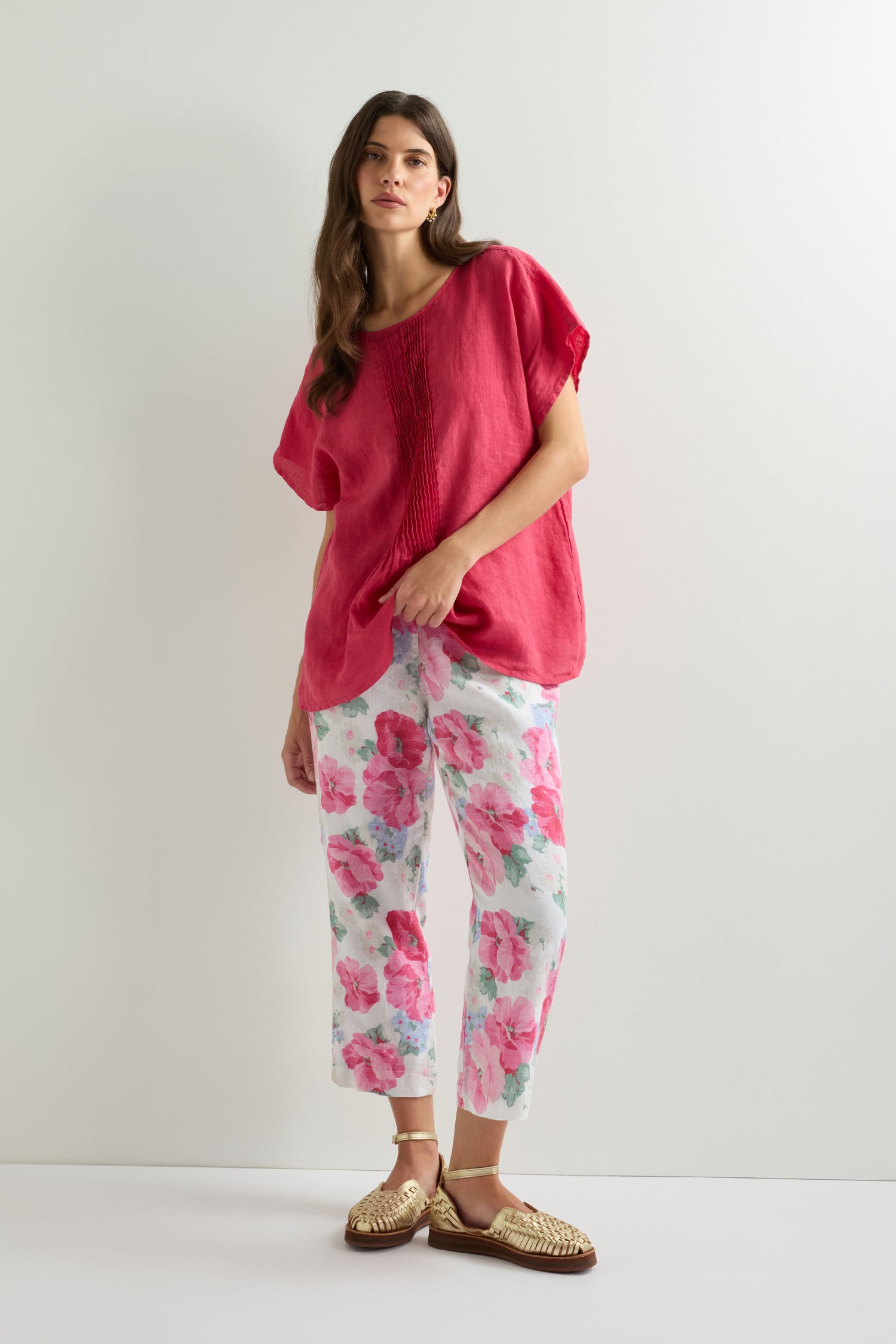 A woman stands against a white wall wearing wide-leg Poppy Print Linen Trousers, a loose red top, and gold woven flats.