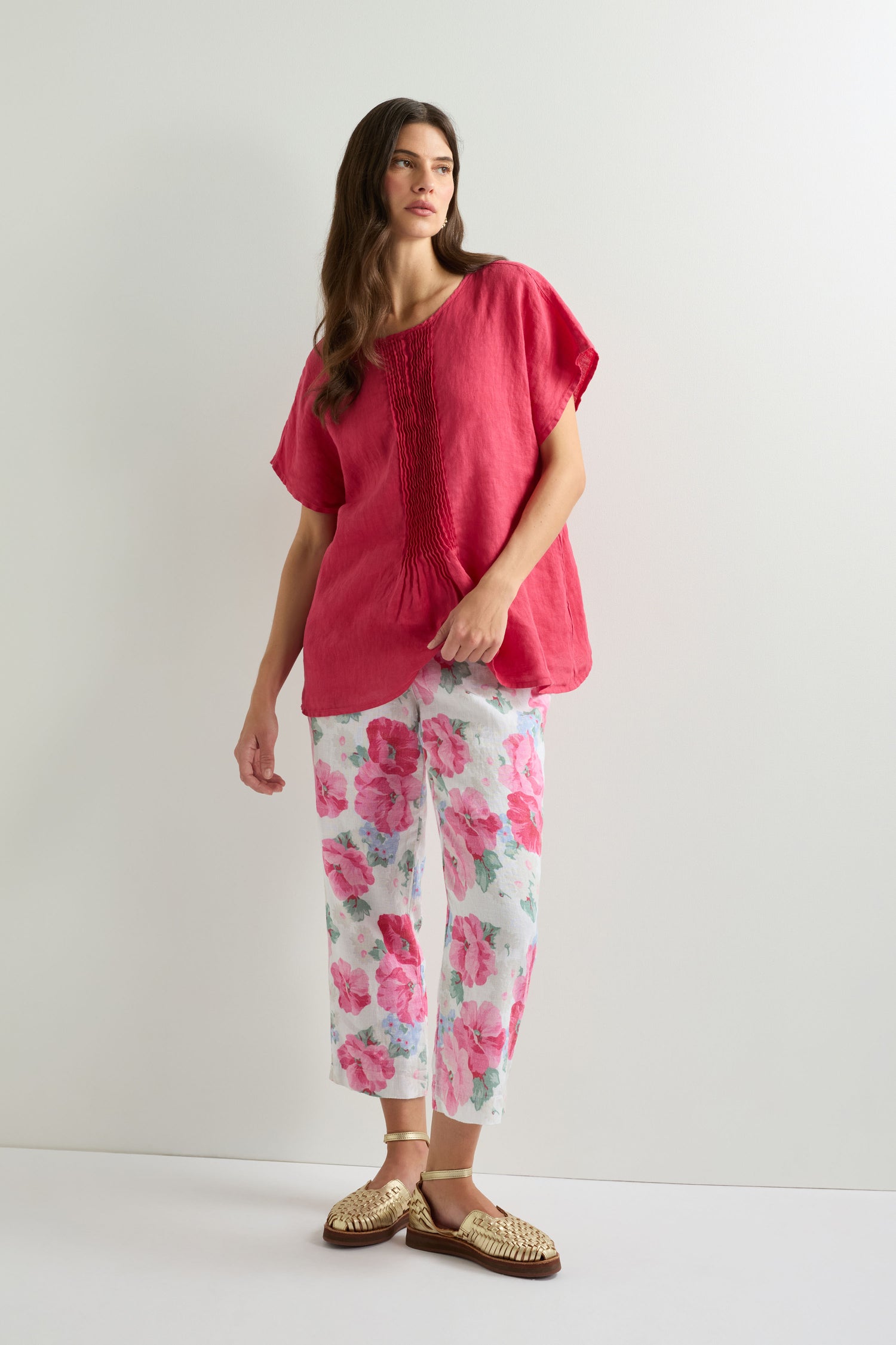 A woman stands against a plain white background, wearing wide-leg Poppy Print Linen Trousers with a loose red top and woven sandals.