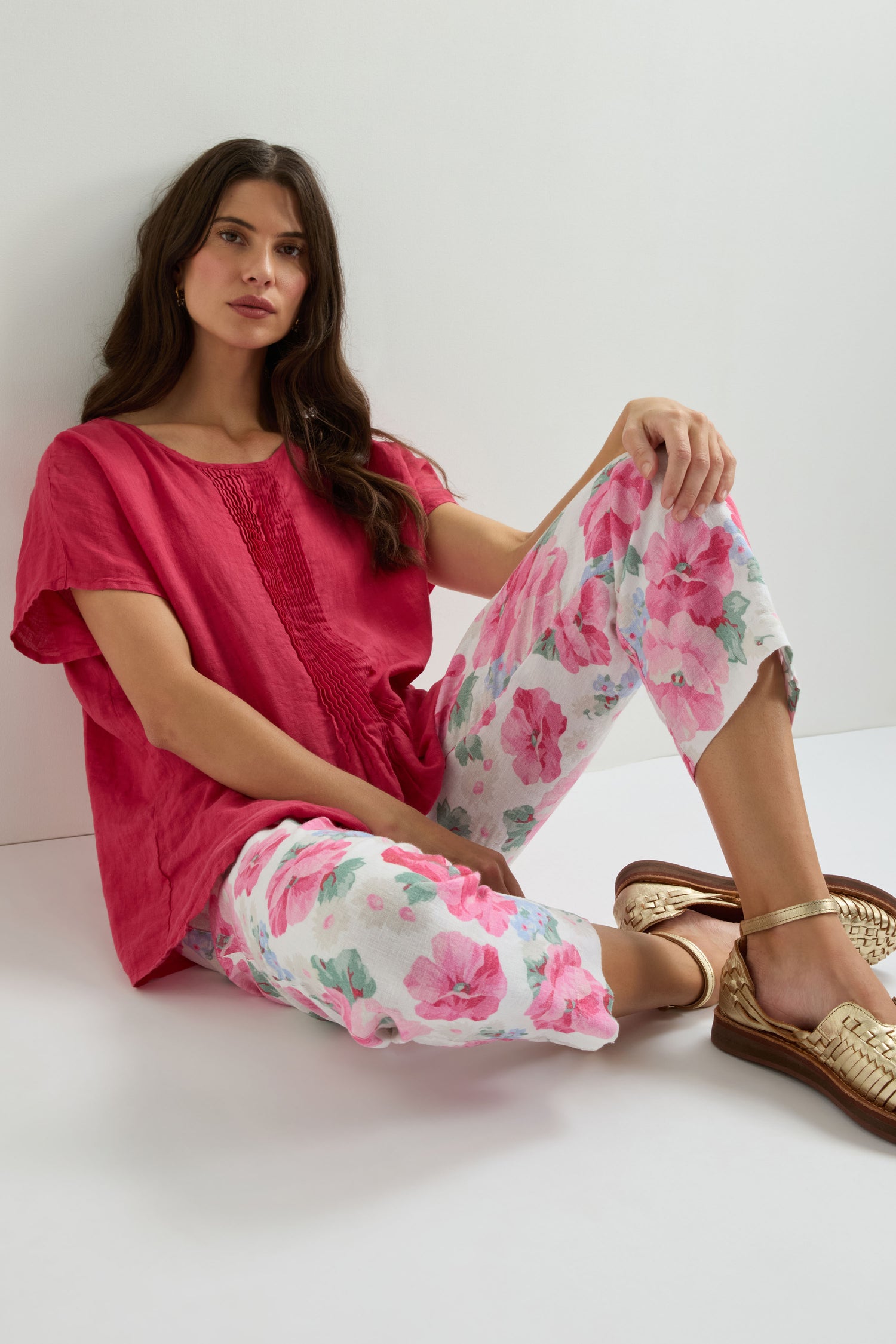 Woman sits against a white wall wearing a loose red top, Poppy Print Linen Trouser featuring a hand-painted pink and white poppy pattern, and gold woven sandals.
