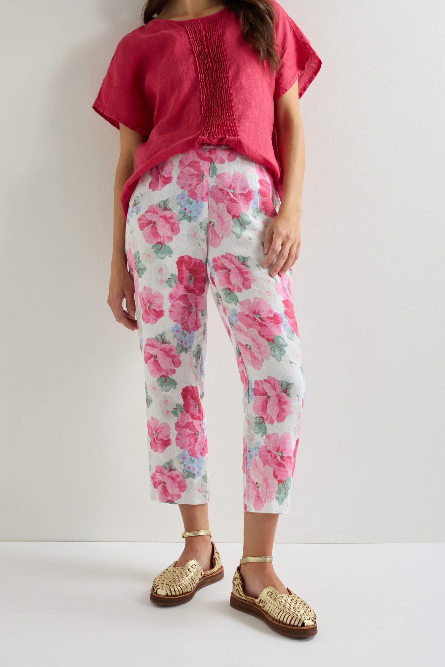 A person stands against a white wall wearing a red short-sleeve top, Poppy Print Linen Trouser with a hand-painted poppy design, and gold woven sandals.