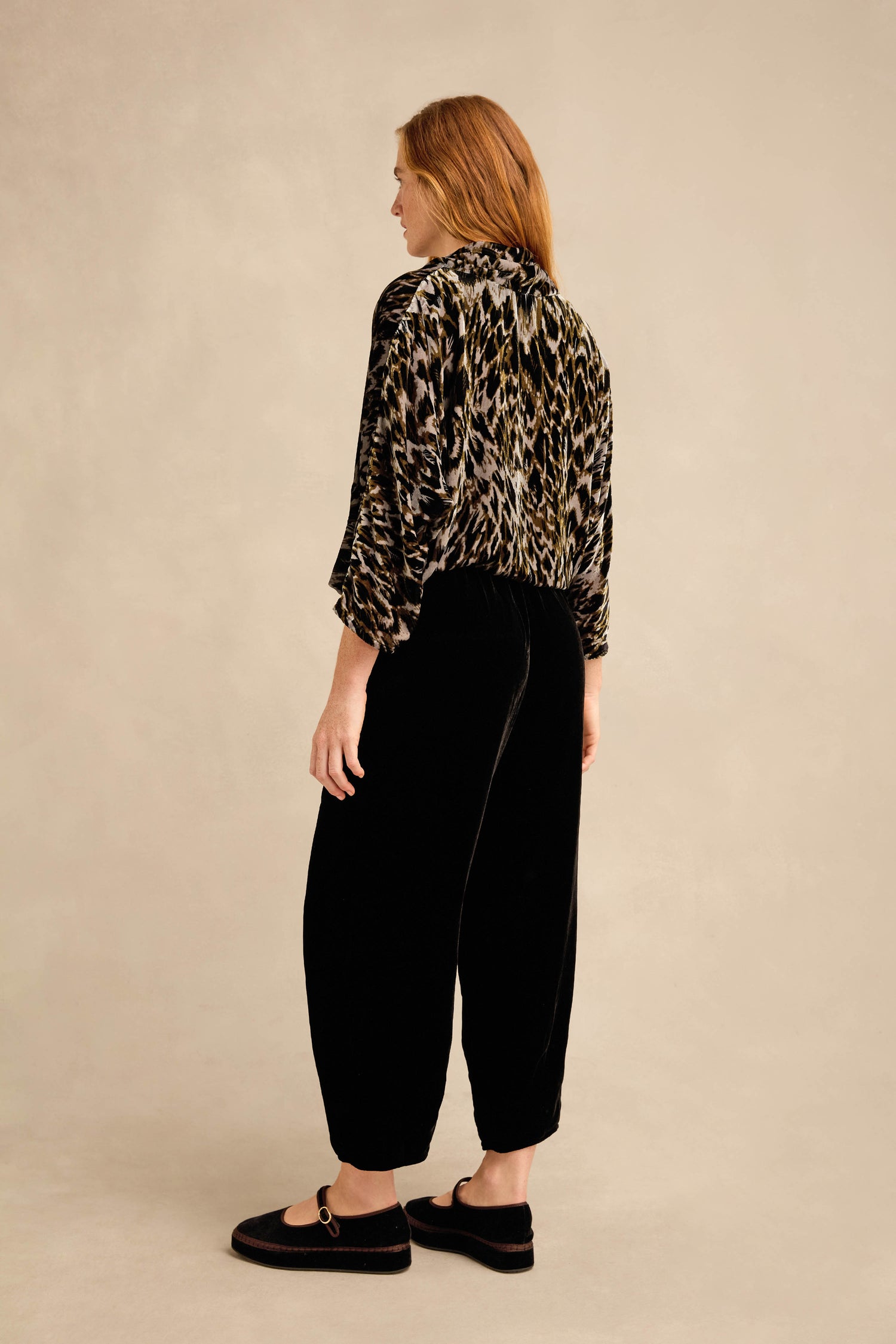 Person standing sideways wearing a leopard print blouse, black silk velvet trousers with a relaxed fit and tapered leg, and black slip-on shoes against a plain beige background.
