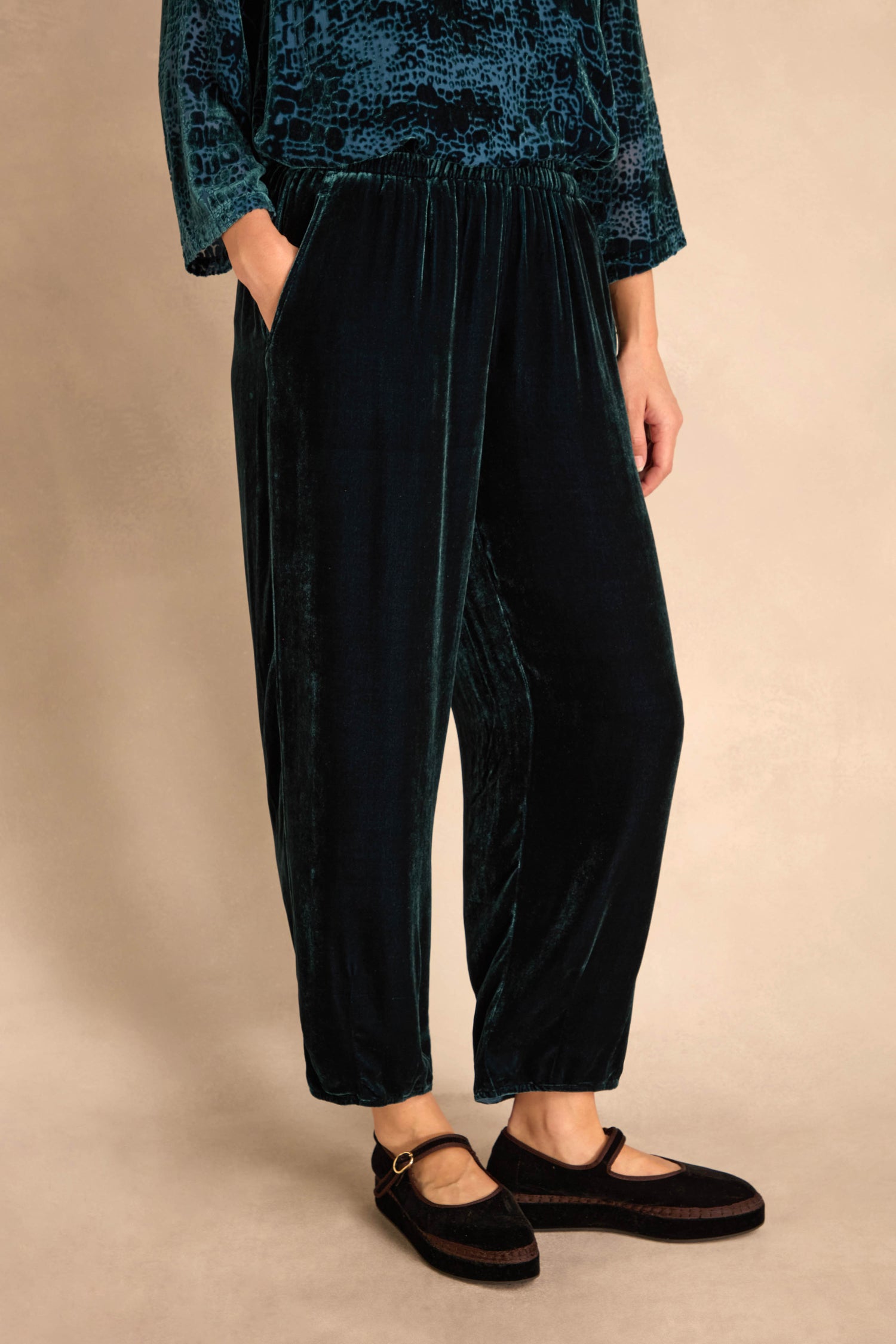 A person in Deep Turquoise Silk Velvet Trousers with an elastic waist, a patterned teal top, and black Mary Jane shoes stands against a neutral background.