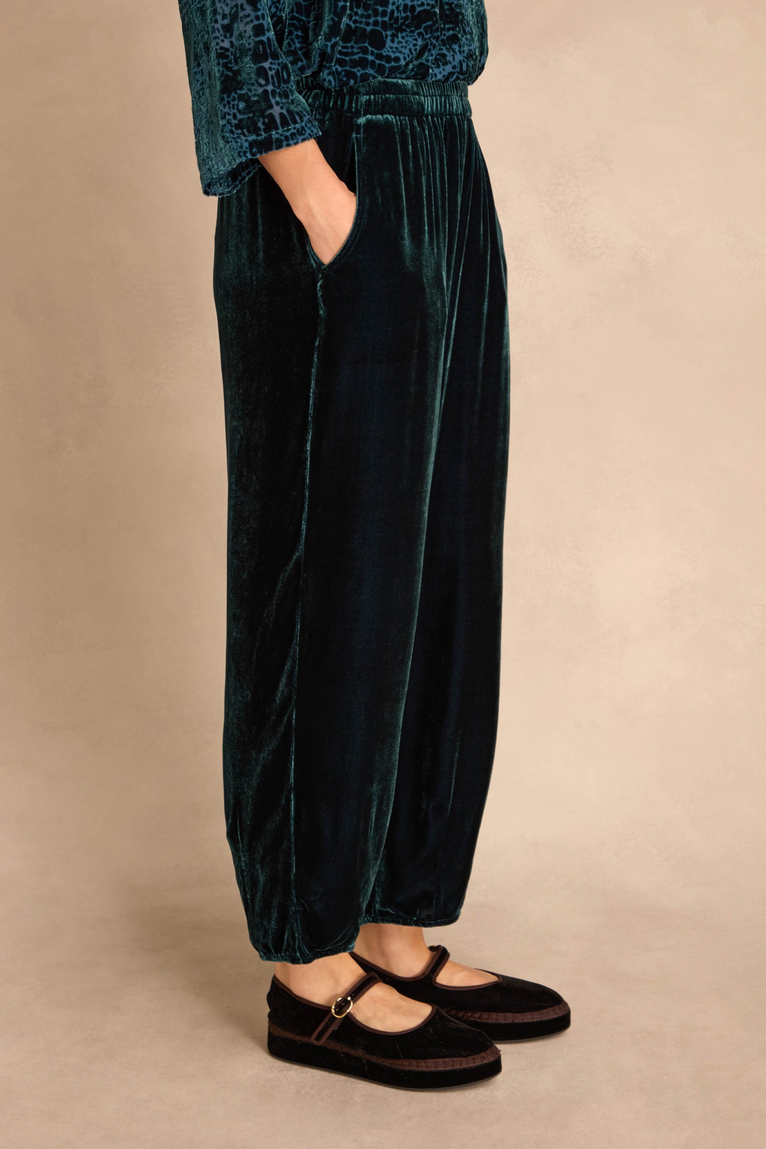 Person wears Deep Turquoise Silk Velvet Trousers with elasticated waist, hands in pockets, and black Mary Jane shoes; shown waist down against a neutral background.