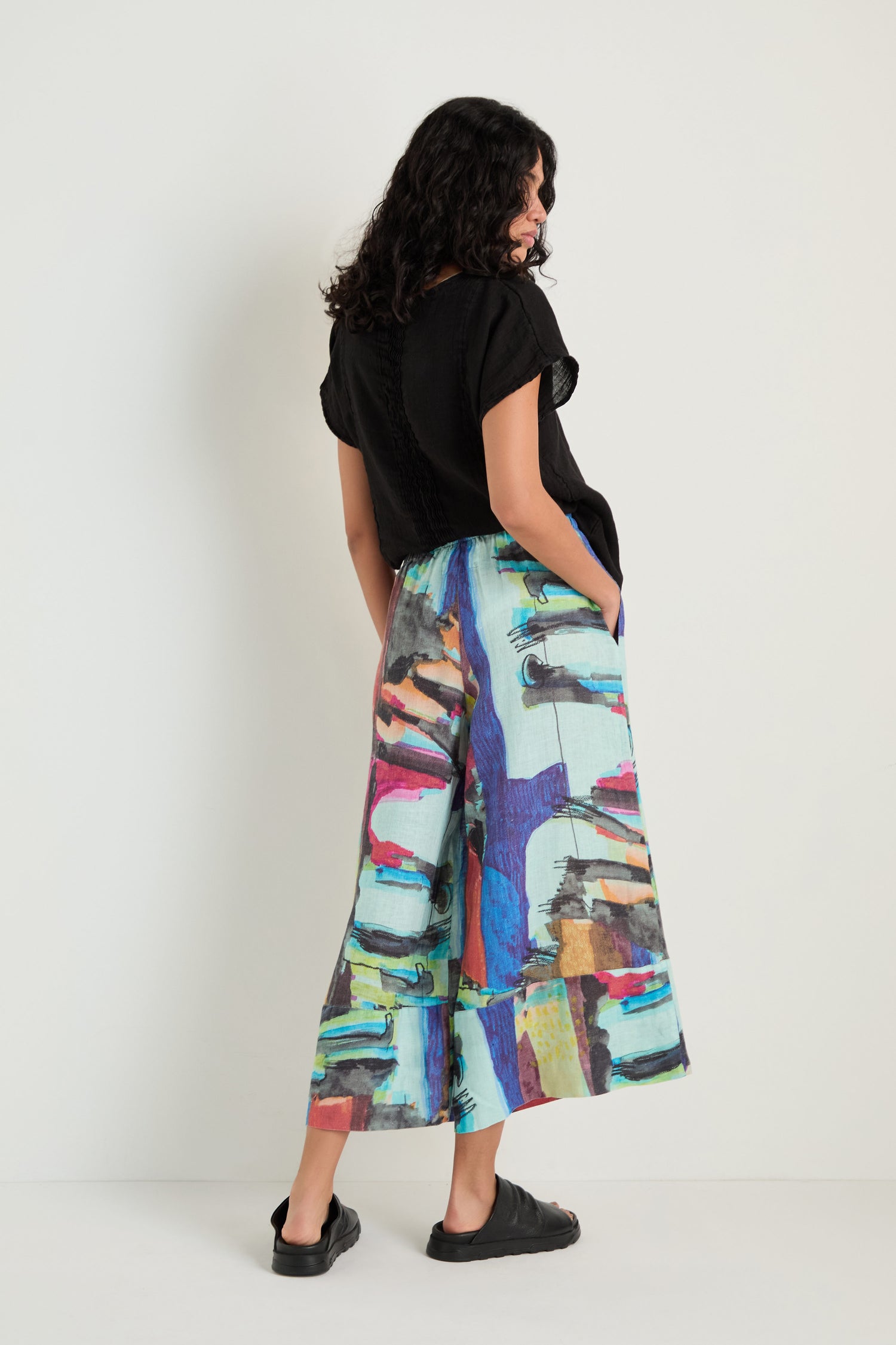 A woman faces away from the camera, hands in her pockets, wearing a black short-sleeved top, black sandals, and Abstract Print Wide Leg Trousers as she stands against a plain wall.