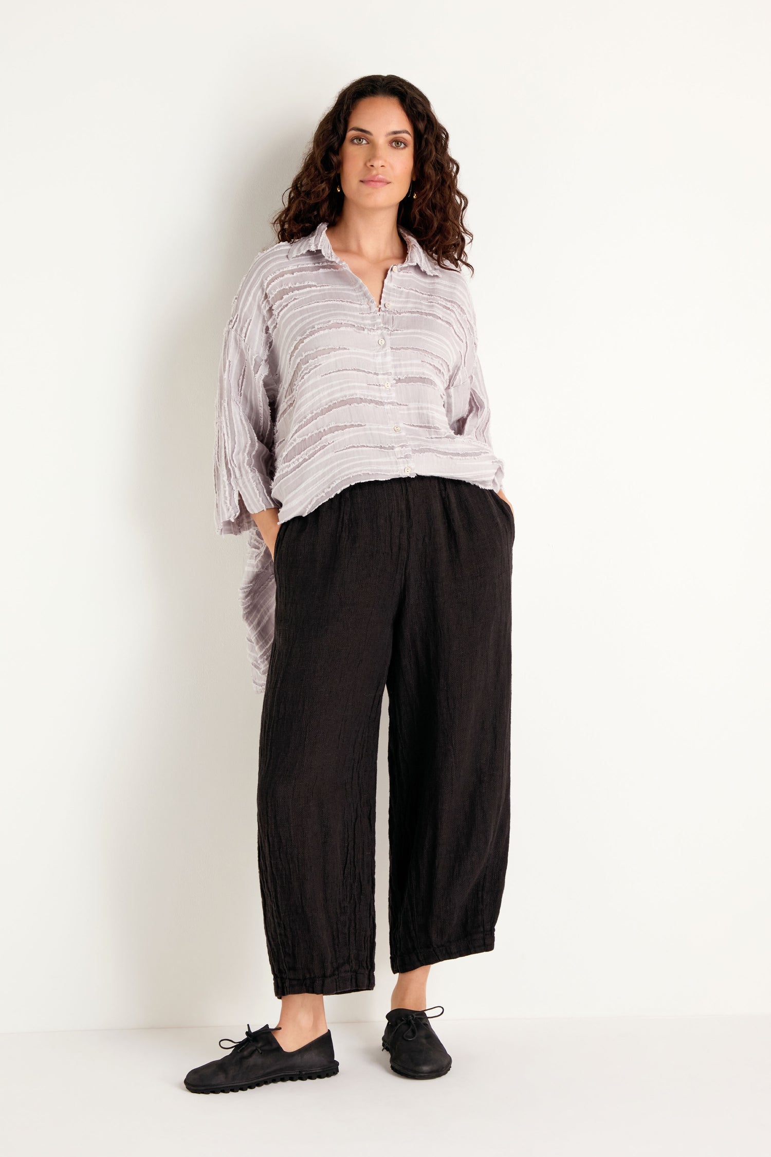 A woman stands against a white background with her hands in her pockets, wearing a loose light striped shirt, black shoes, and the Textured Linen Charcoal Bubble Trousers made from breathable natural fabric.