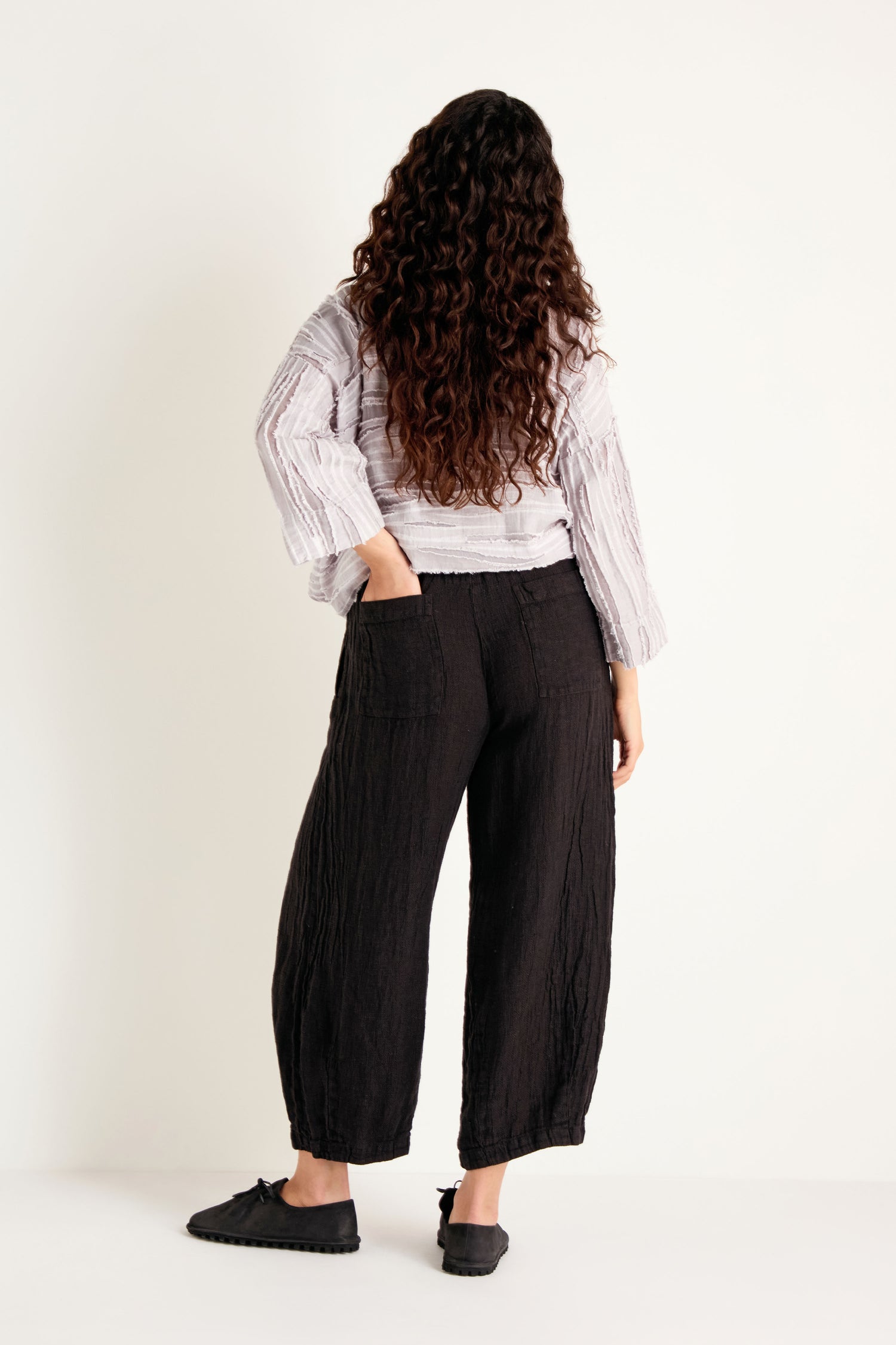 A person with long curly hair stands facing away, wearing a textured light long-sleeve top, the Textured Linen Charcoal Bubble Trousers crafted from breathable natural fabric, and black shoes, with one hand in a pocket, against a plain white background.