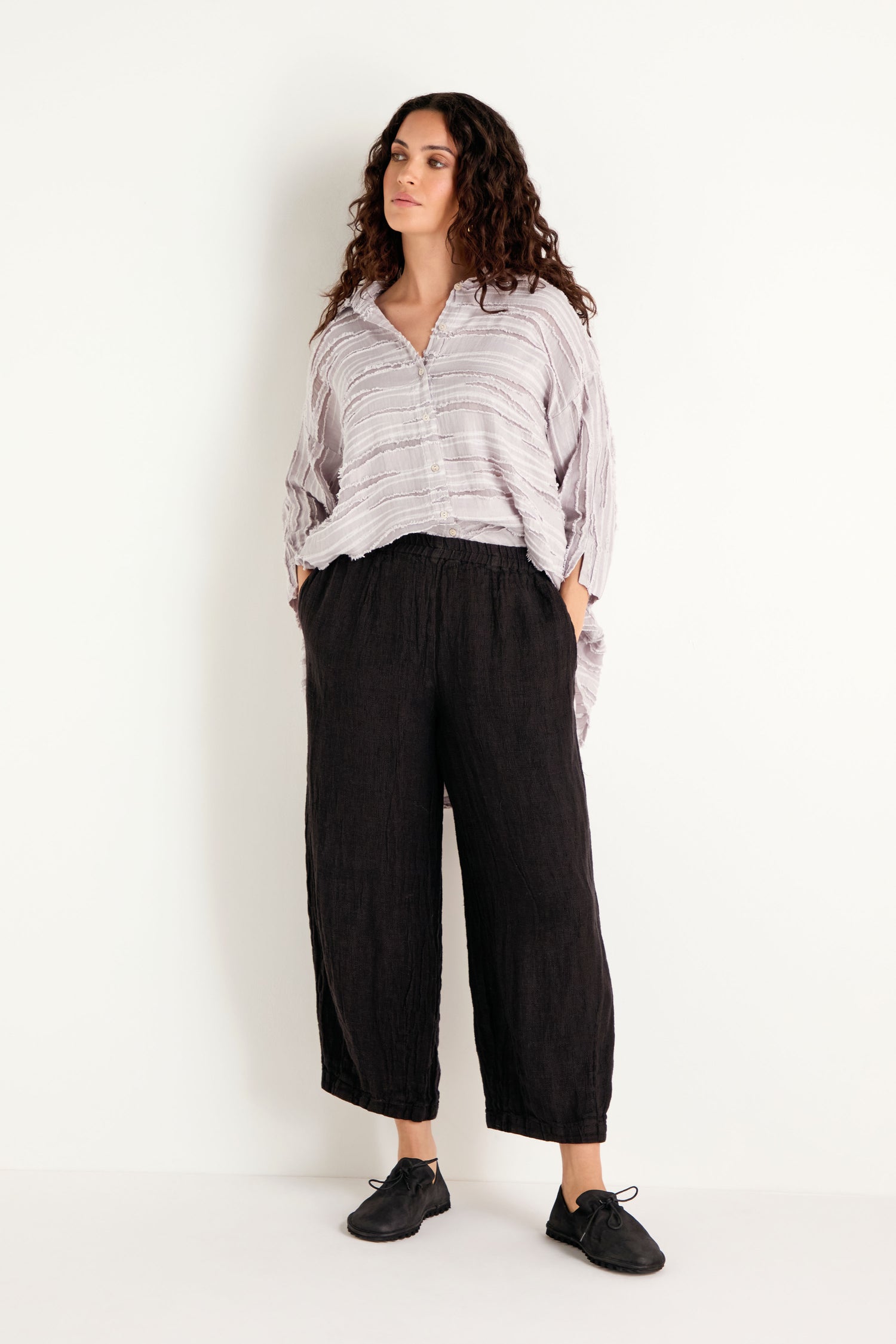 A woman stands against a white wall wearing a loose striped blouse and Textured Linen Charcoal Bubble Trousers crafted from breathable natural fabric, paired with black shoes, hands in pockets and looking to the side.