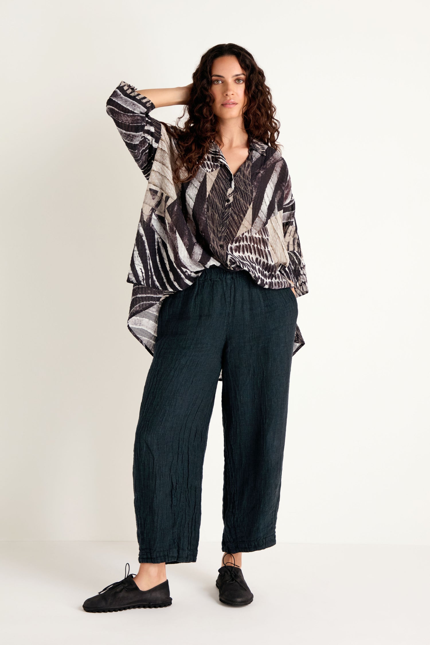 A woman stands against a white background wearing a patterned long-sleeve top, Textured Linen Turquoise Bubble Trousers, and black shoes.