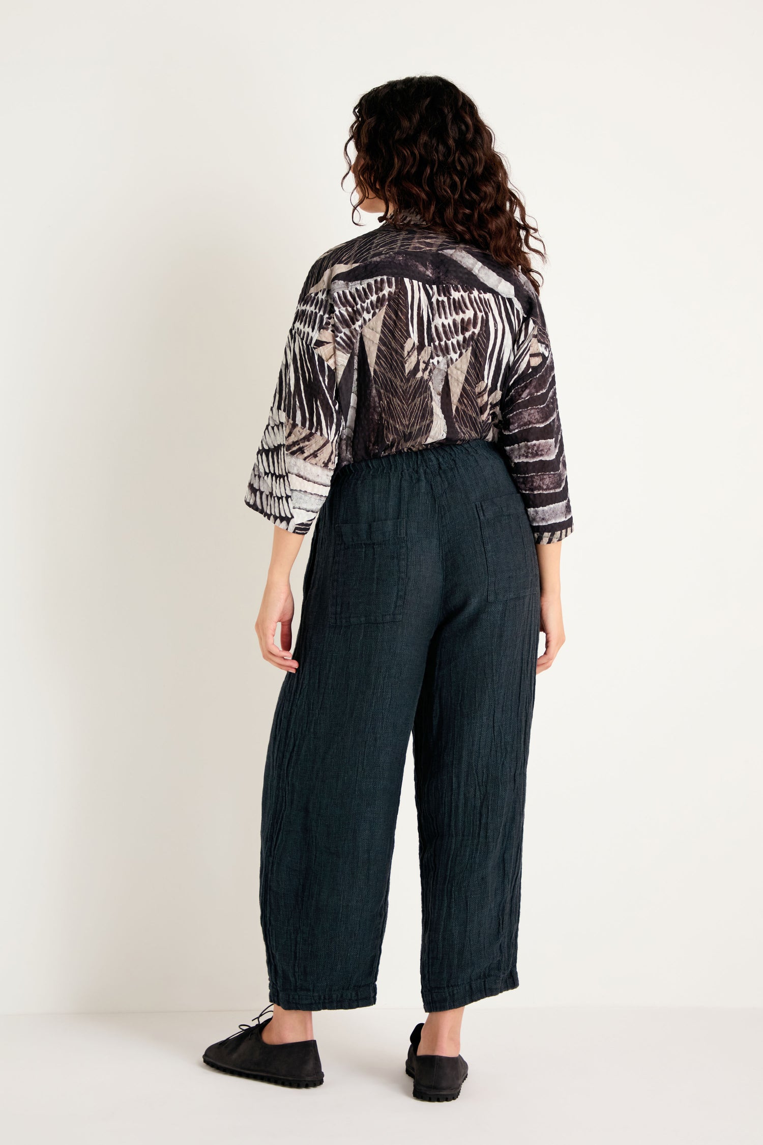 A person stands facing away, dressed in a black and white patterned top paired with Textured Linen Turquoise Bubble Trousers and black shoes against a plain light background.
