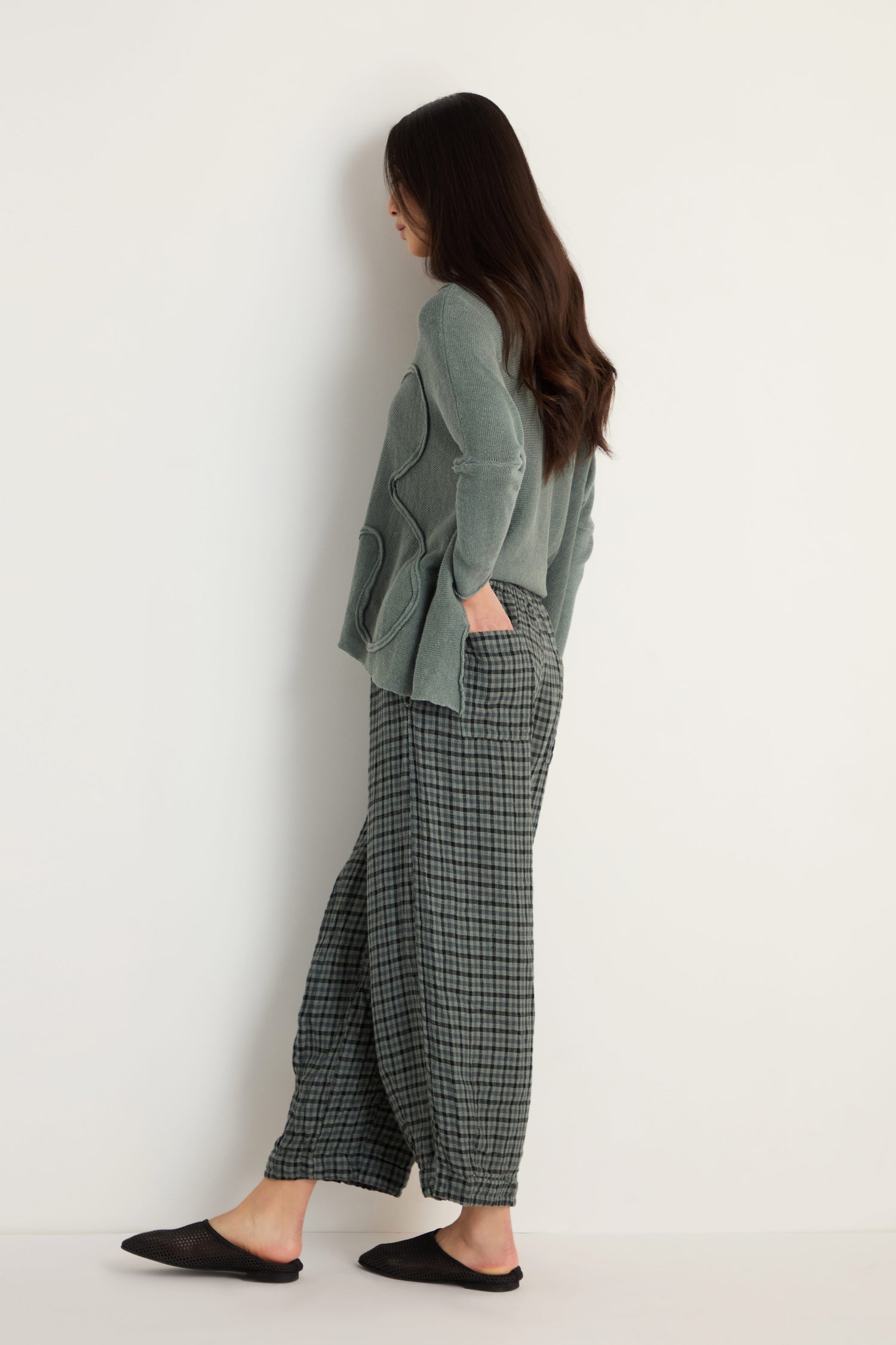 A person stands sideways against a plain white wall, wearing Check Linen Bubble Trousers with a relaxed silhouette, a sage green sweater, and black slip-on shoes.
