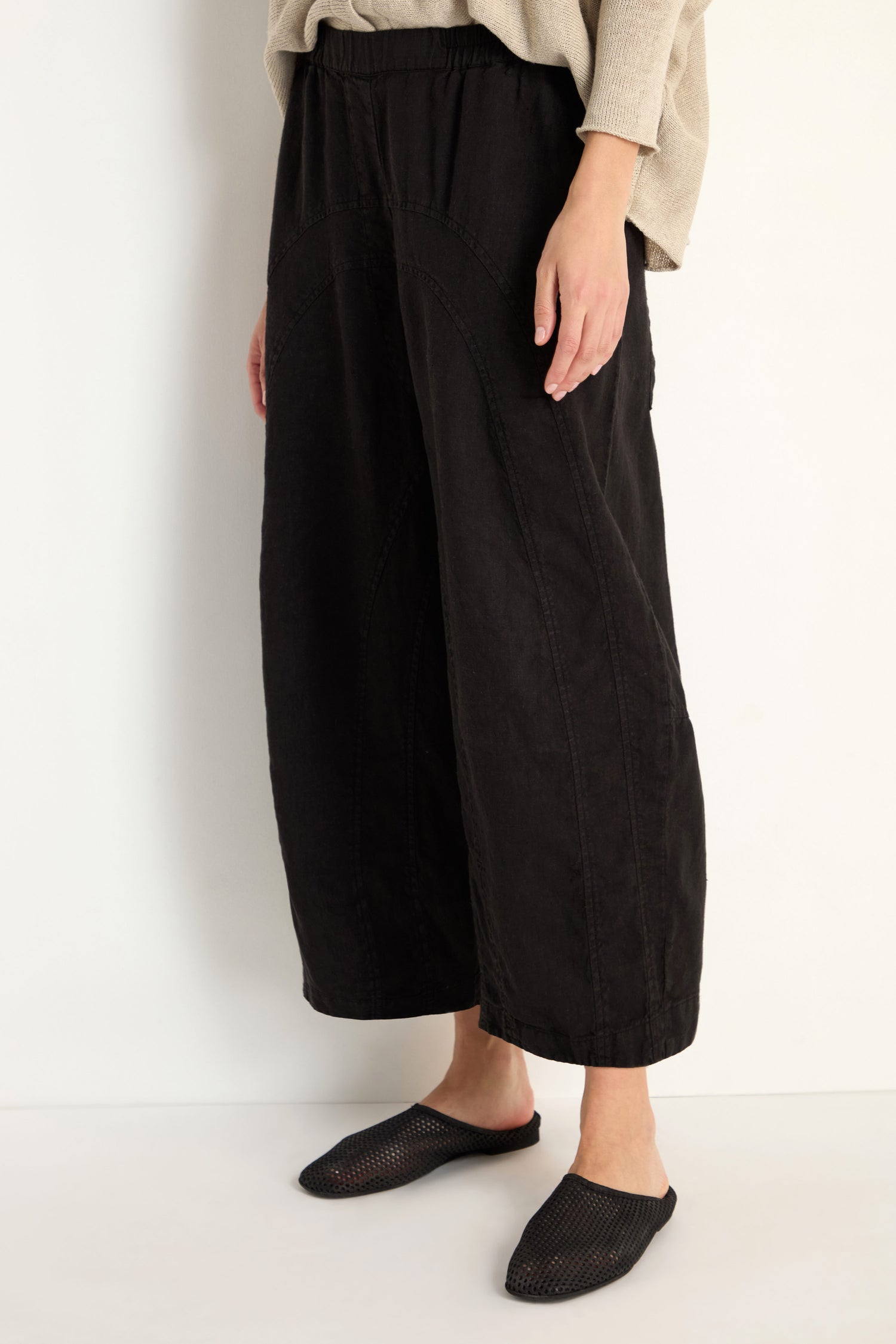 Model wears Curved Seam Linen Trousers in black with a relaxed leg, paired with a beige top and black slip-on shoes, against a plain white background.