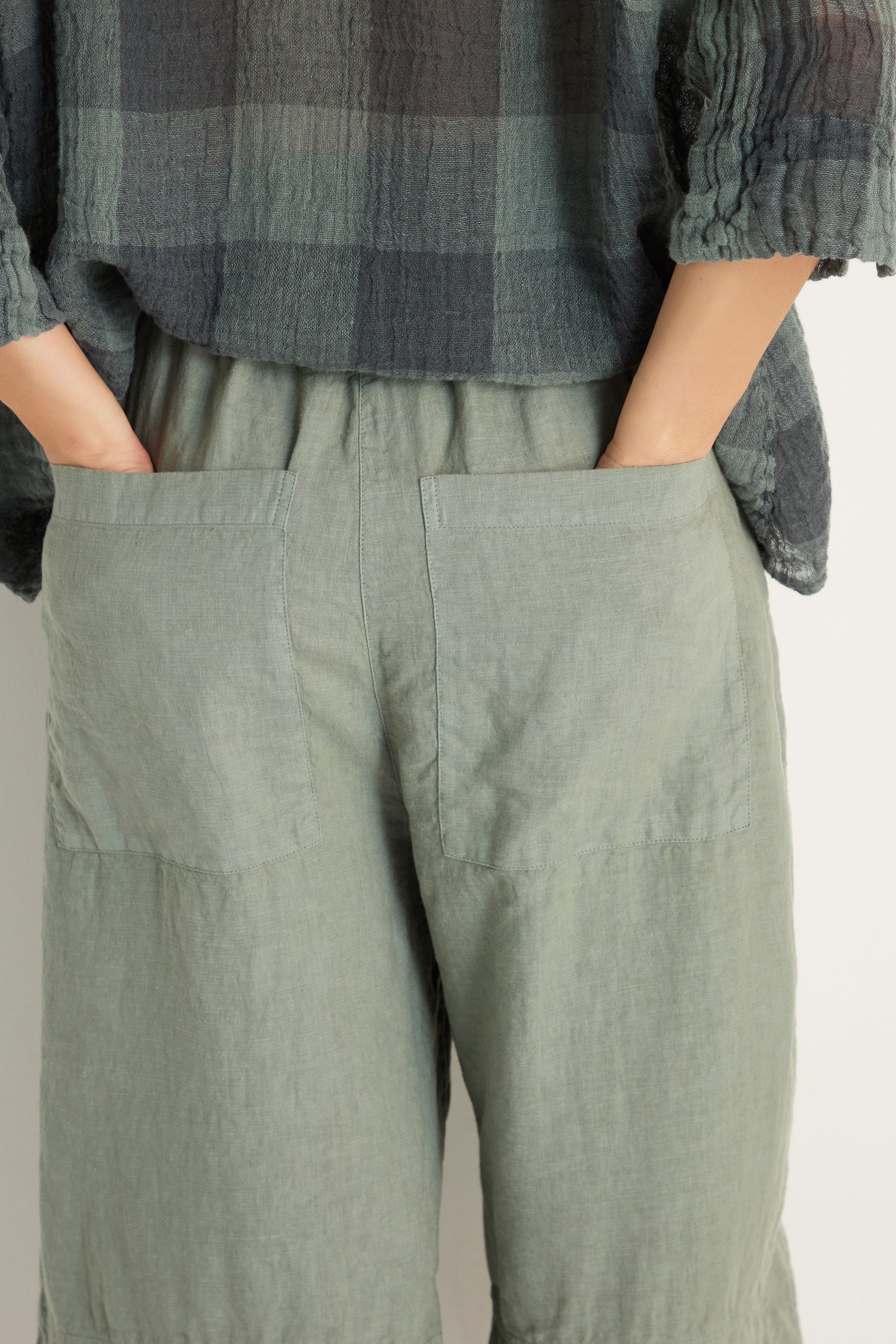 Person photographed from behind wearing Curved Seam Linen Trousers in light green, paired with a loose dark plaid shirt, hands tucked into the back pockets.