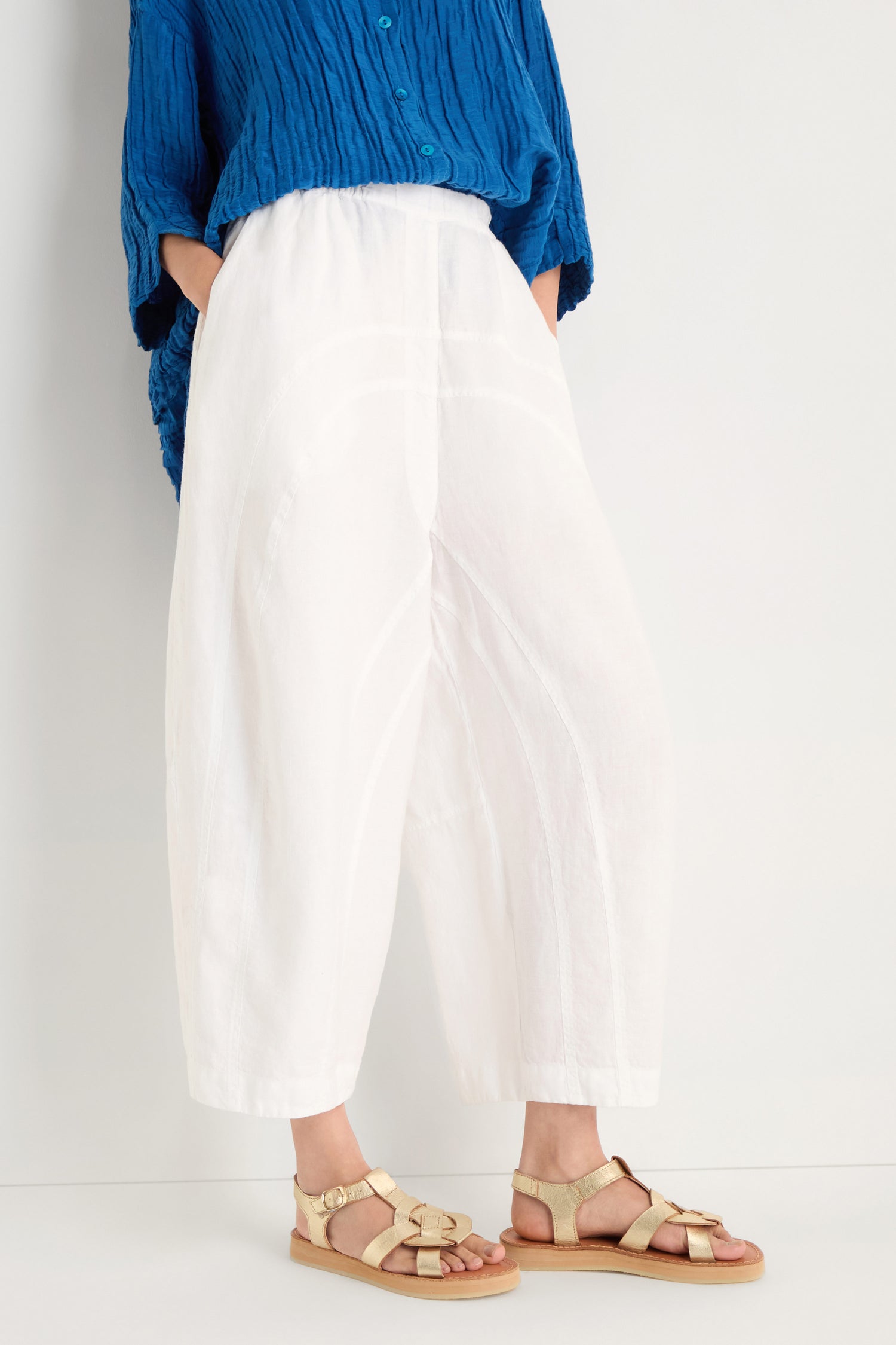 Person wearing a blue textured shirt, Curved Seam White Linen Trousers featuring a relaxed silhouette, and beige strappy sandals, standing against a plain light background.