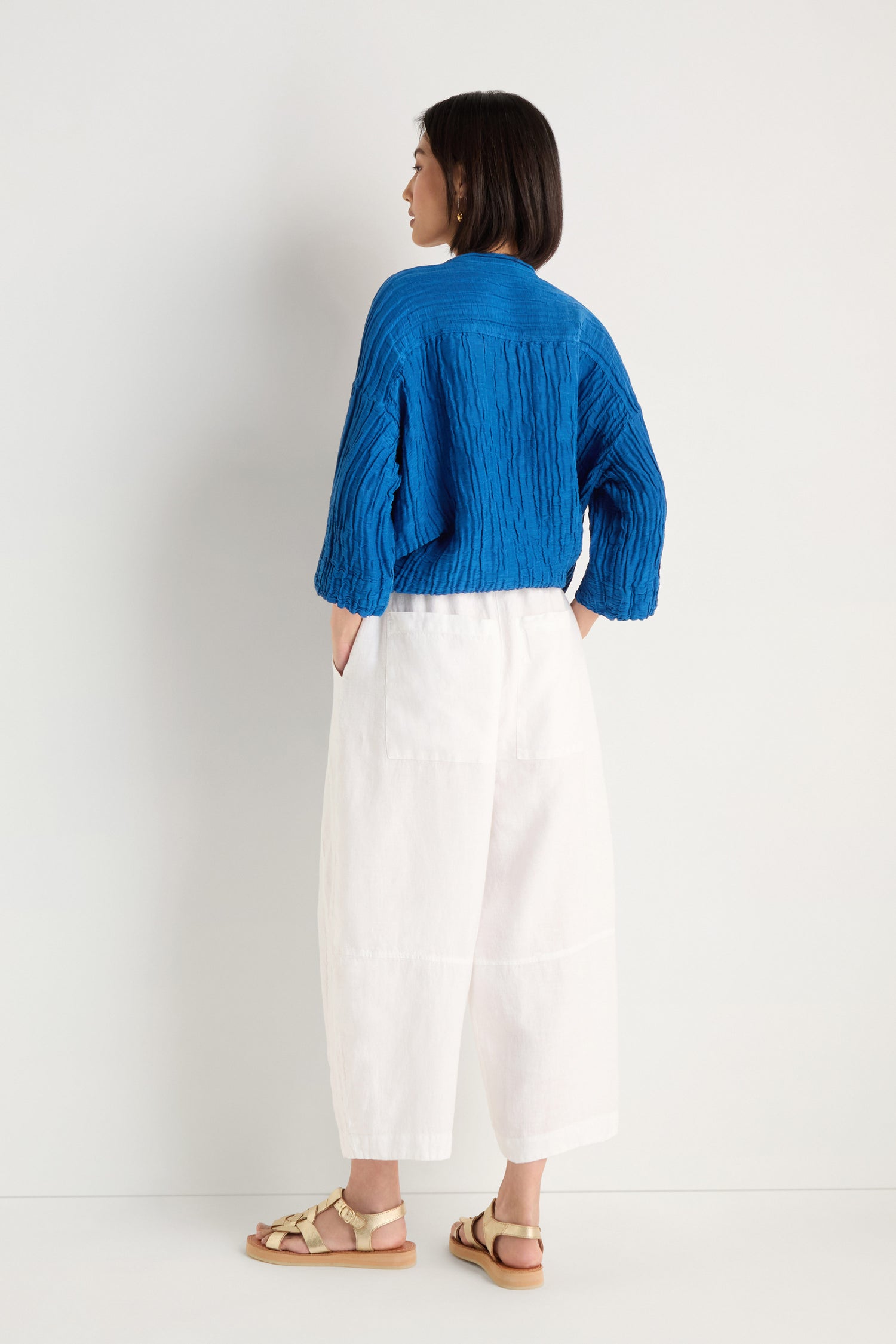 A person stands facing away, wearing a textured blue long-sleeve top, Curved Seam White Linen Trousers with a relaxed fit, and beige sandals against a plain white background.