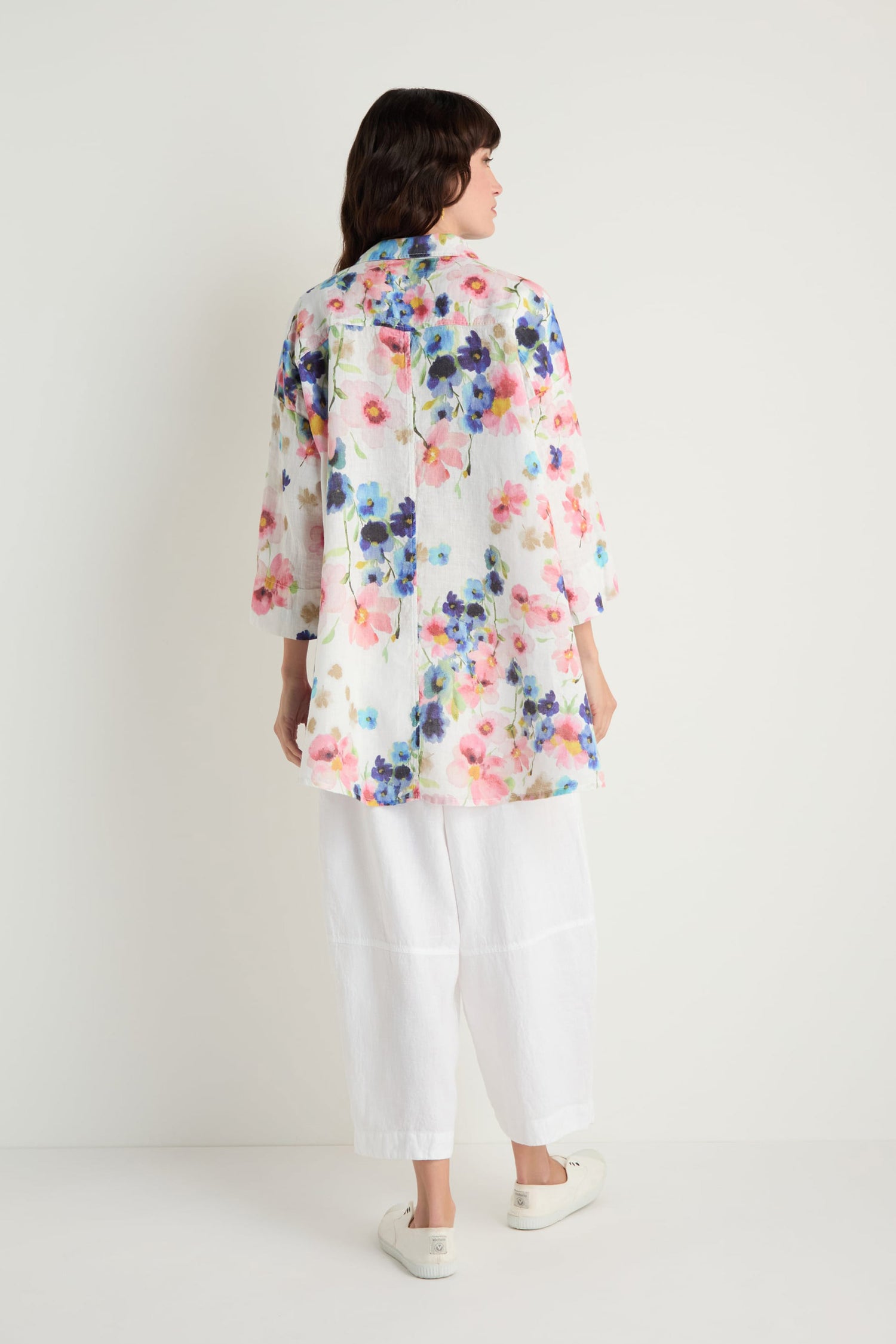A person stands facing away, wearing the Primrose Print Flared Linen Shirt, paired with white wide-leg pants and slip-on shoes against a plain light background.