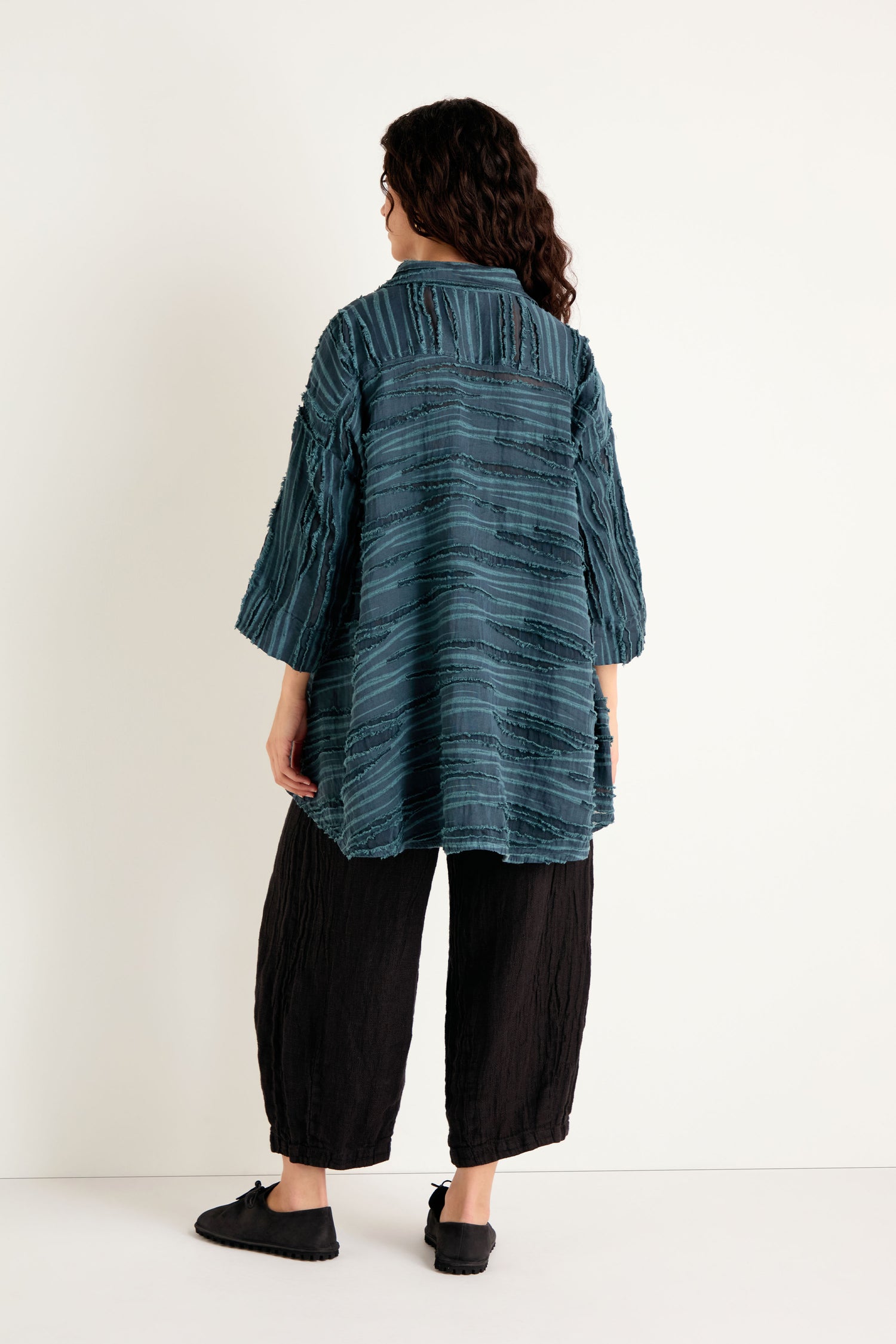 A person is standing against a white wall, facing away from the camera, wearing a Deep Turquoise Textured Stripe Flared Shirt, loose black pants, and black shoes.